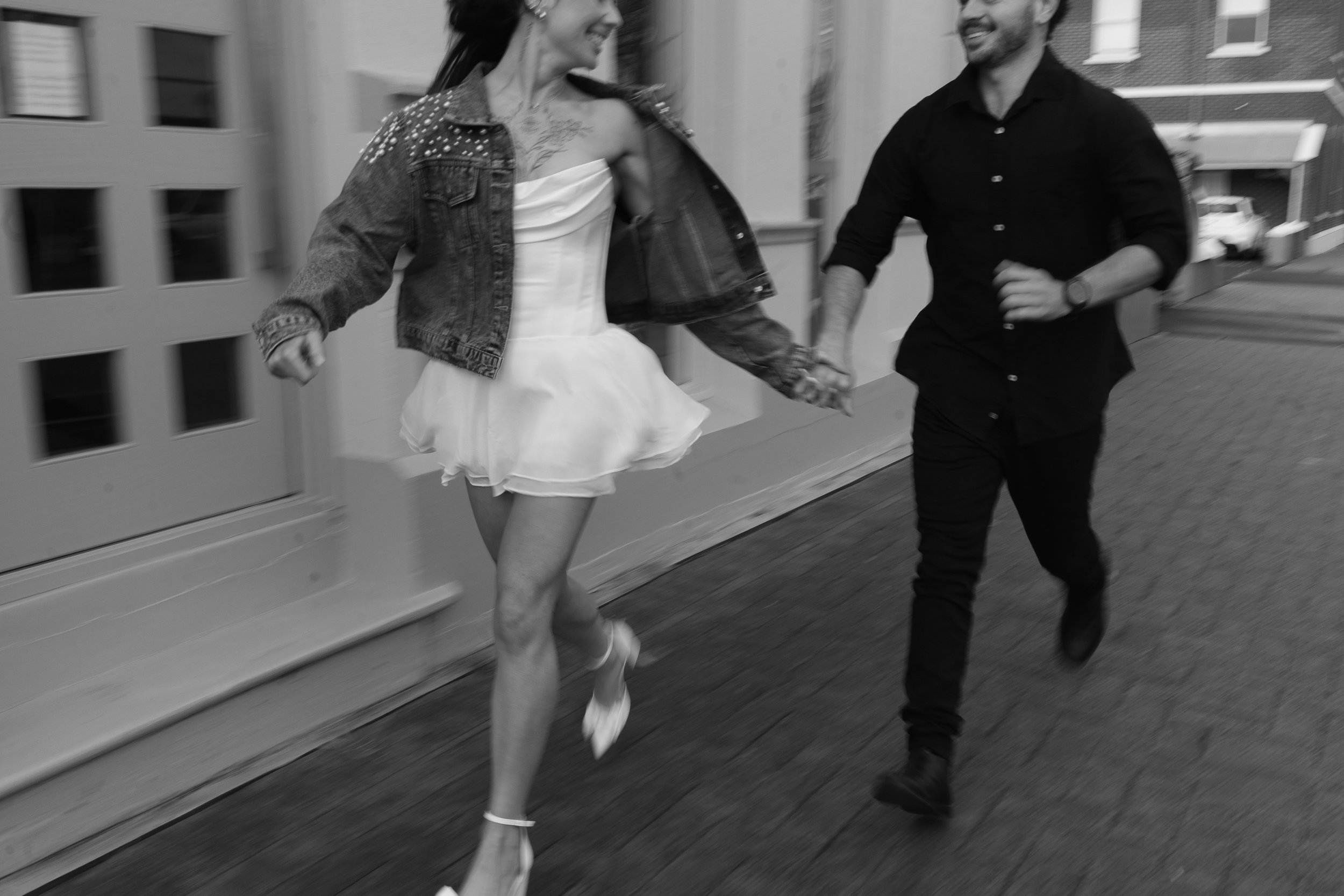 A woman and a man holding hands and running on a city street, woman wearing a short white dress and denim jacket, man wearing black shirt and pants, both smiling.
