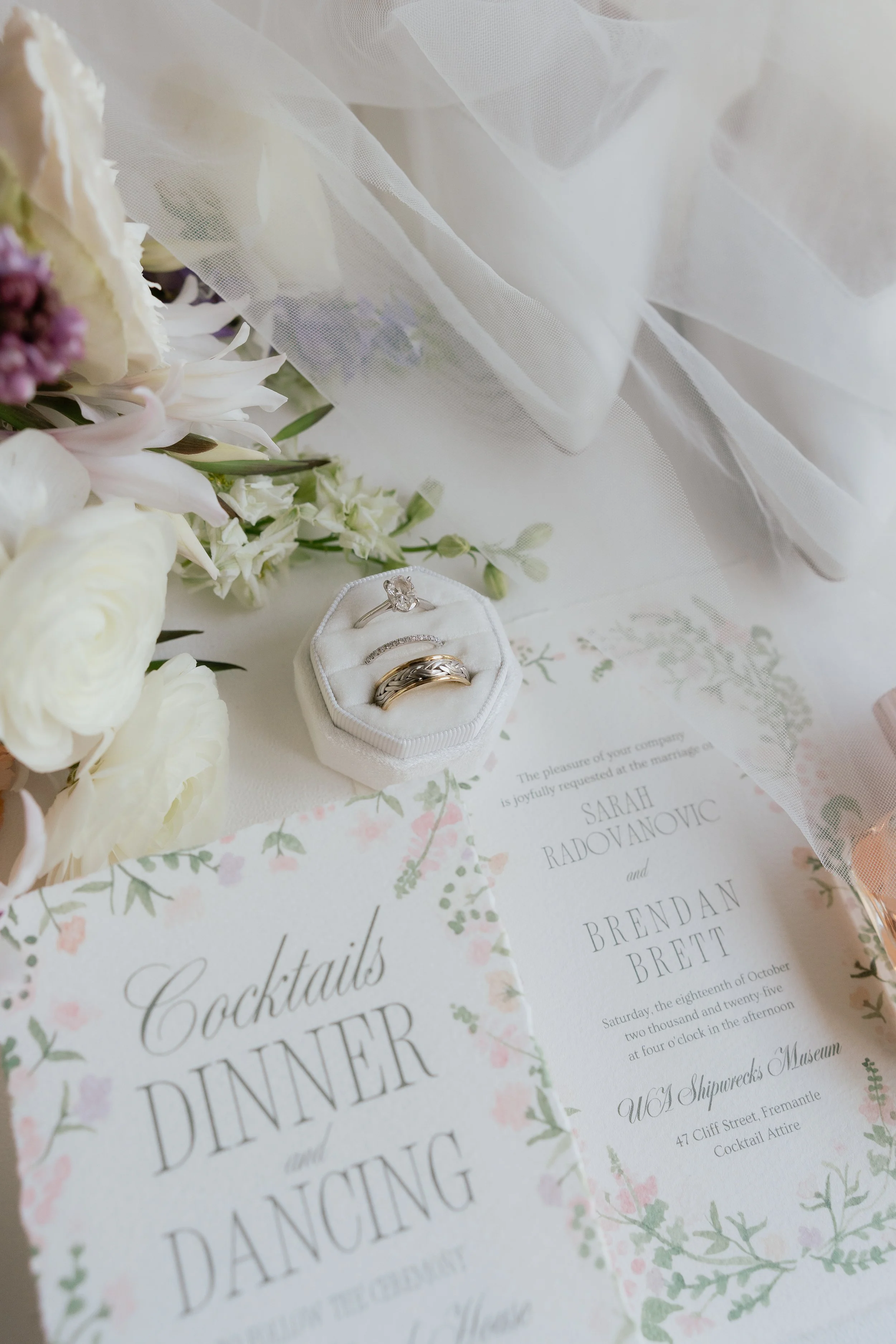 Wedding rings on a white cushion surrounded by flowers, with wedding invitations nearby, set on a delicate, semi-transparent white veil.