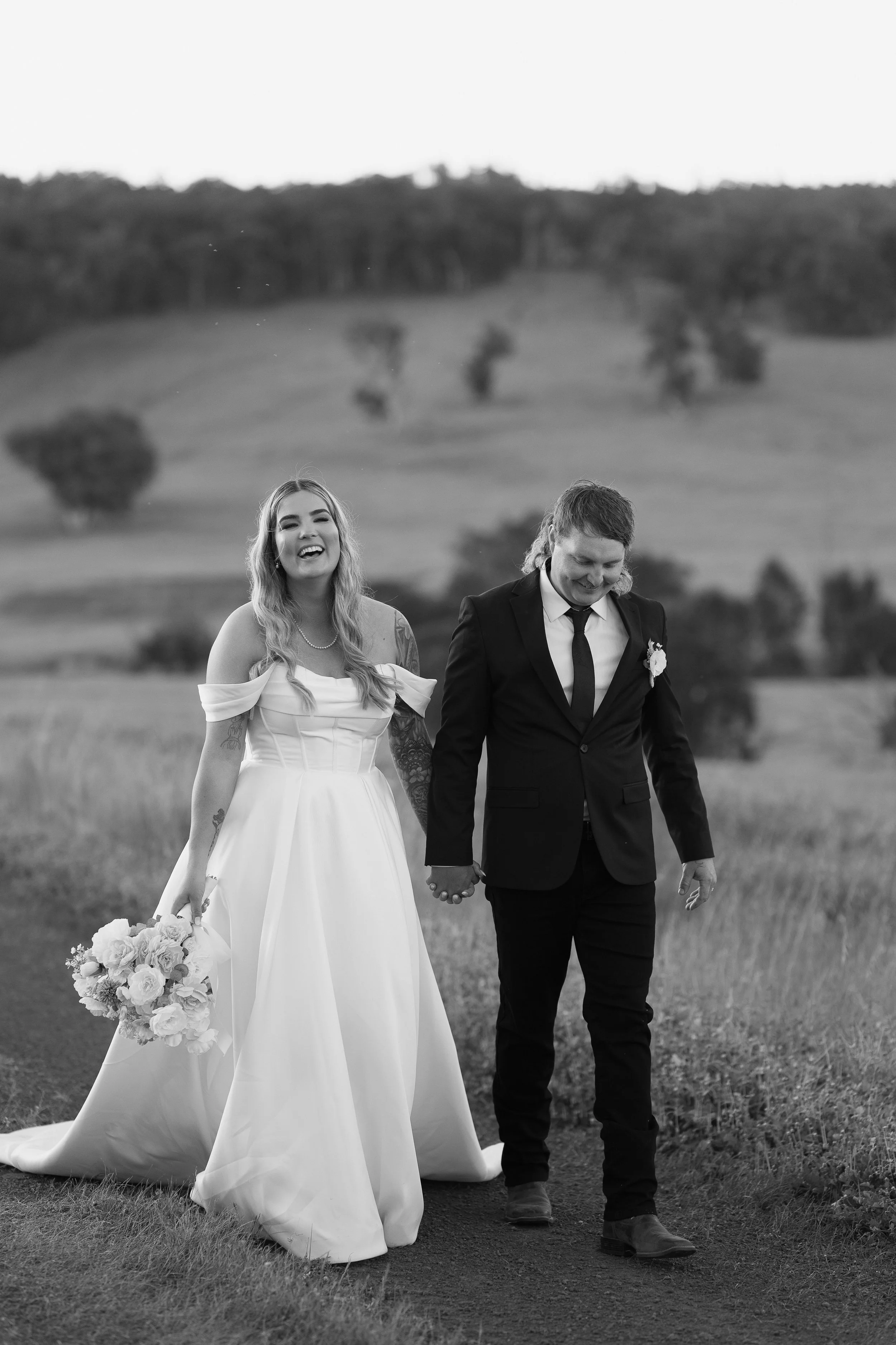 A black and white photo of a happy couple walking hand in hand outdoors in a field, with rolling hills and scattered trees in the background. The woman wears a white wedding dress and holds a bouquet, while the man wears a black suit with a white shi