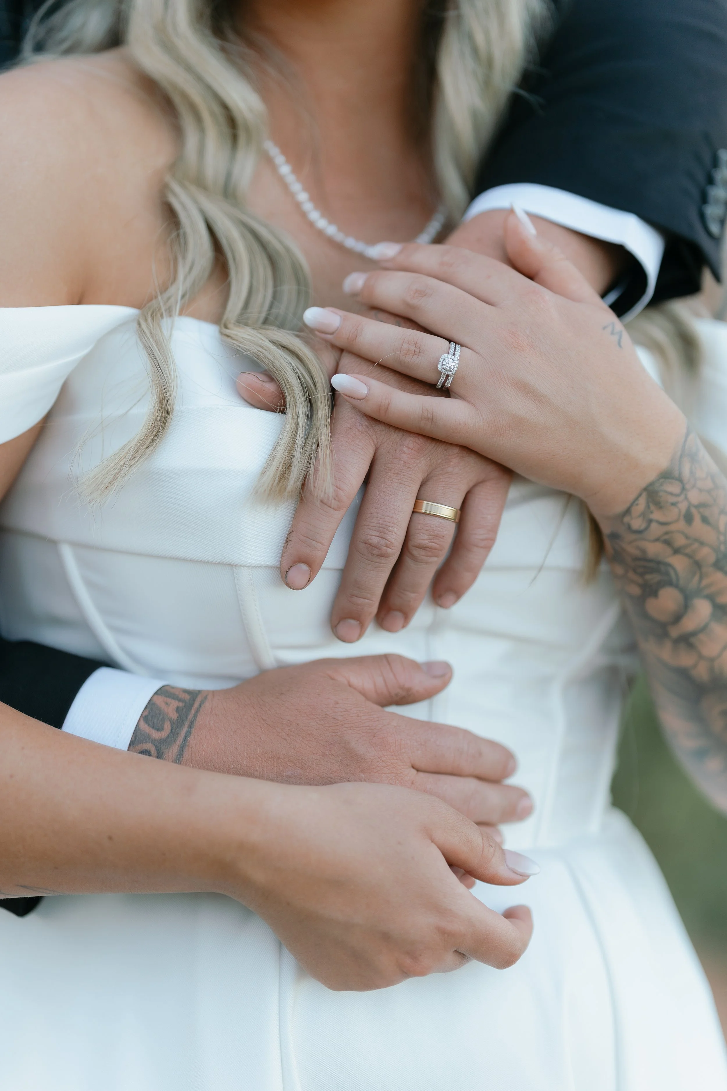 Close-up of a bride and groom holding their hands over her chest, showing wedding rings. The bride has blonde hair, a pearl necklace, and a tattooed arm. The groom has a tattooed arm and is wearing a black suit.