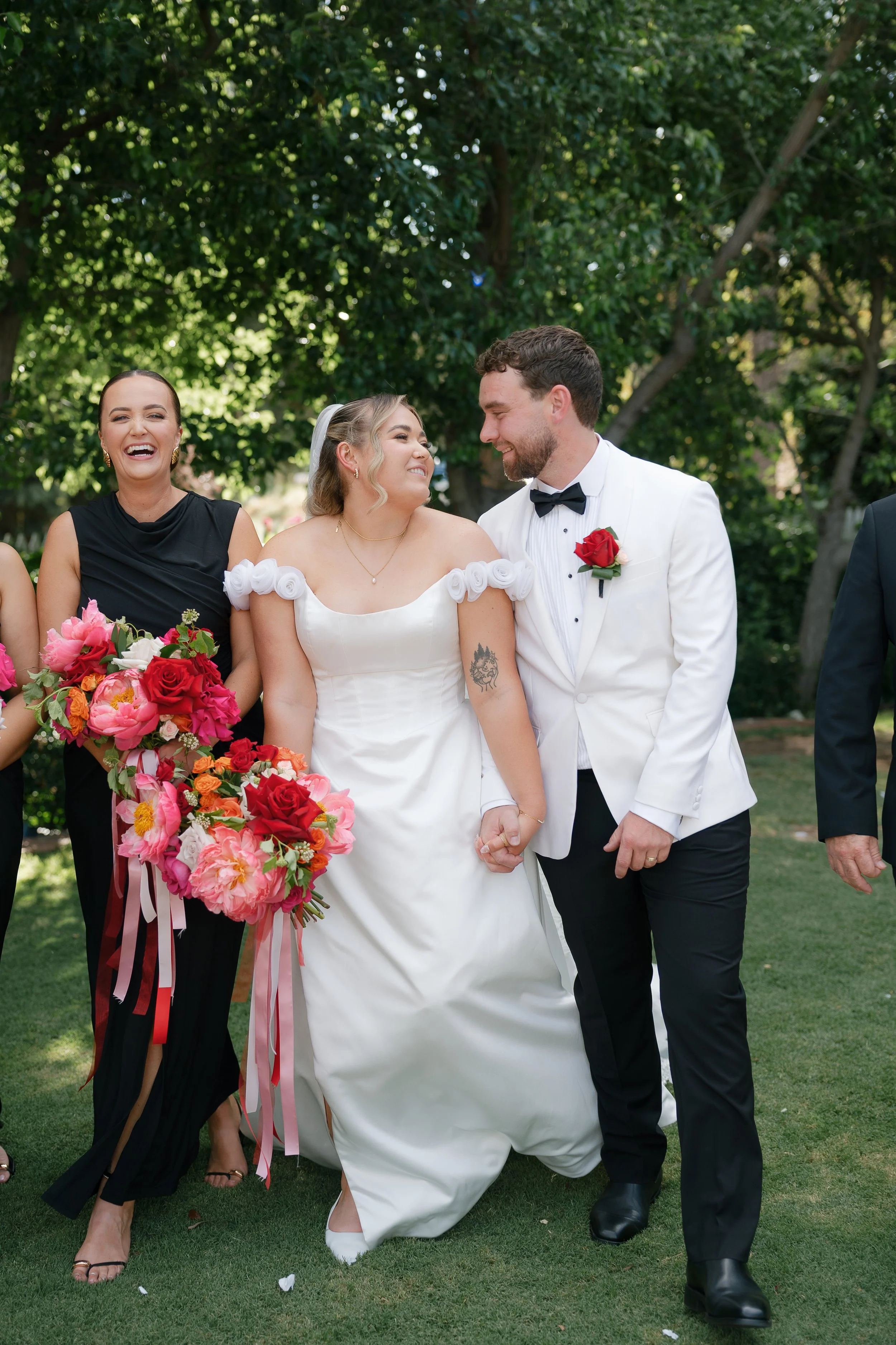 A wedding scene with a bride, groom, and a bridesmaid outdoors. The bride is smiling and holding a bouquet of pink and red flowers. The groom, dressed in a white tuxedo, is holding the bride's hand and smiling. The bridesmaid, in a black dress, stand
