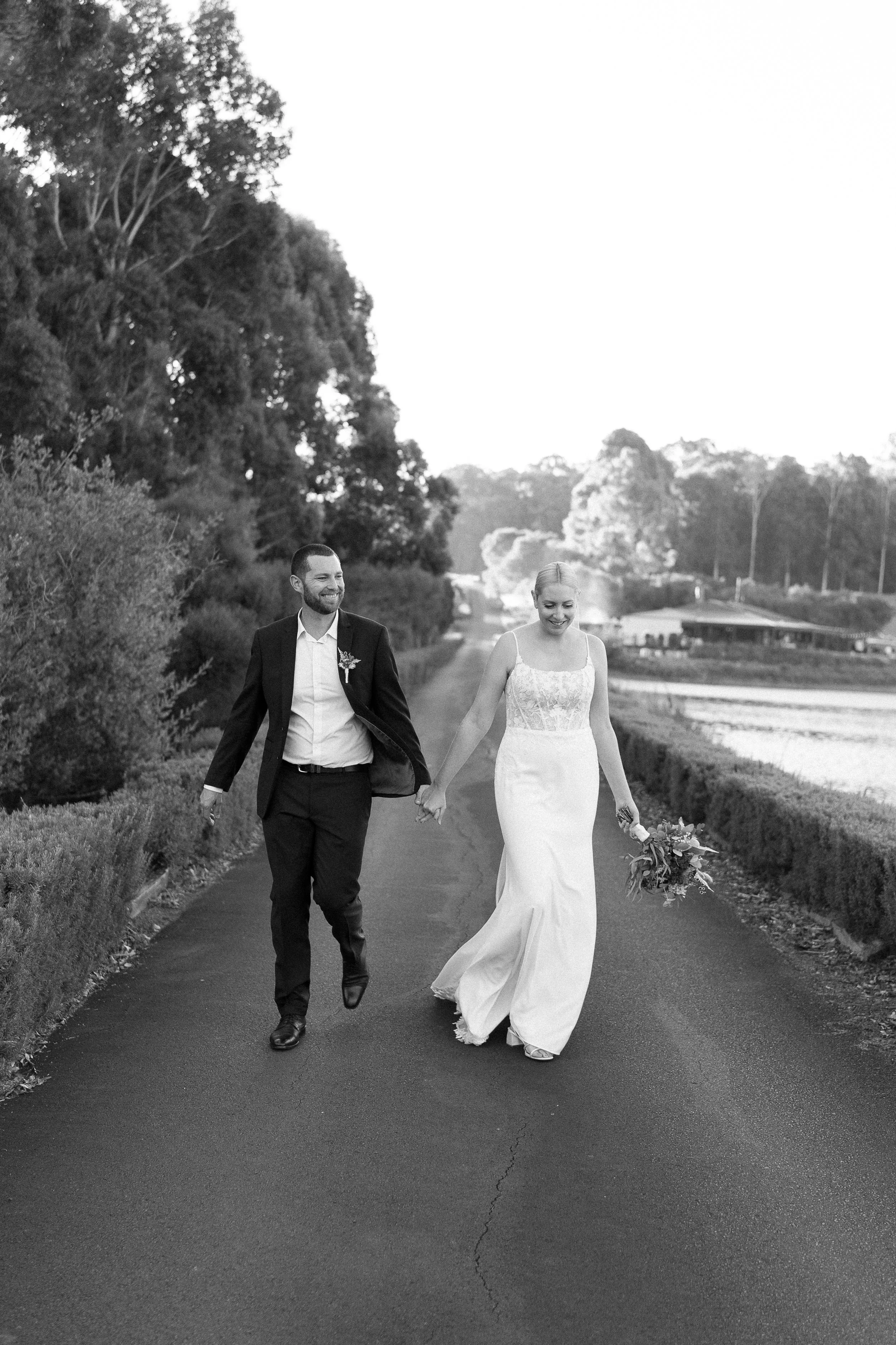 A bride and groom walking hand in hand along a rural road. The bride is wearing a white wedding dress and holding a bouquet, while the groom is dressed in a suit, both smiling and looking joyful. The scene is outdoors with trees on one side and an op