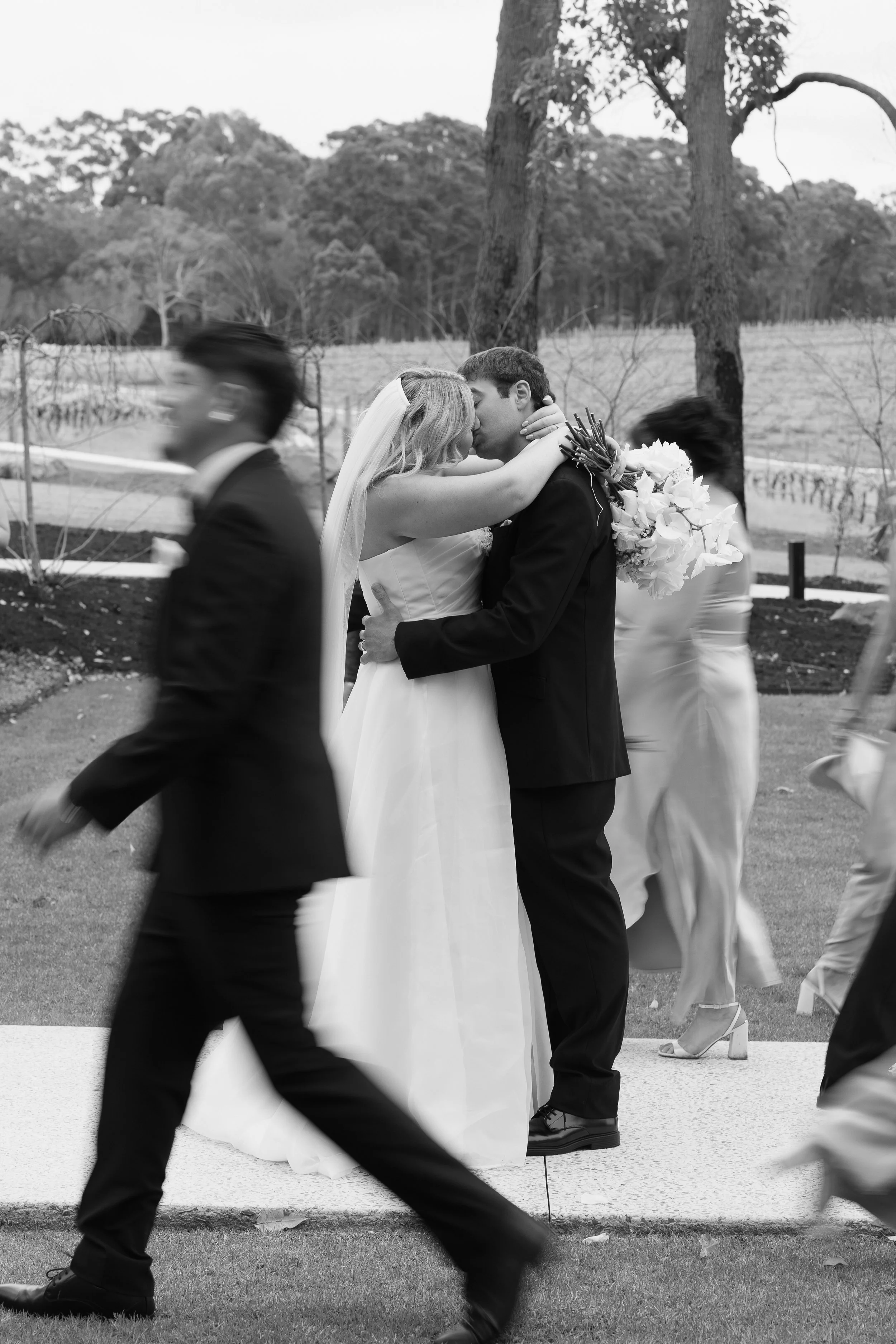 A black-and-white photo of a wedding scene with a bride and groom kissing, surrounded by moving guests, outdoors with trees and landscape in the background.