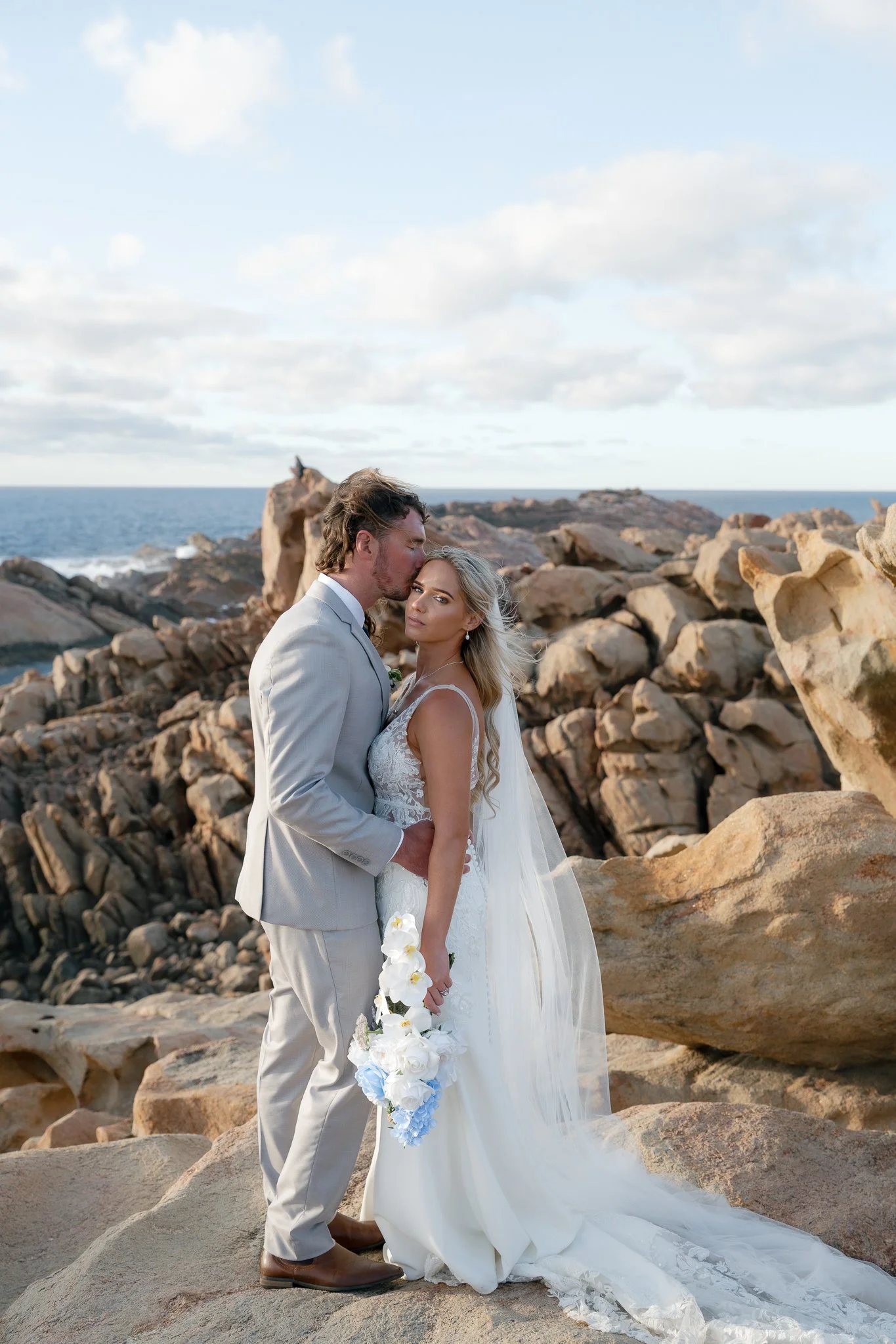 A couple in wedding attire standing on rocks by the ocean, with the groom kissing the bride's forehead and the bride holding a bouquet of white flowers.