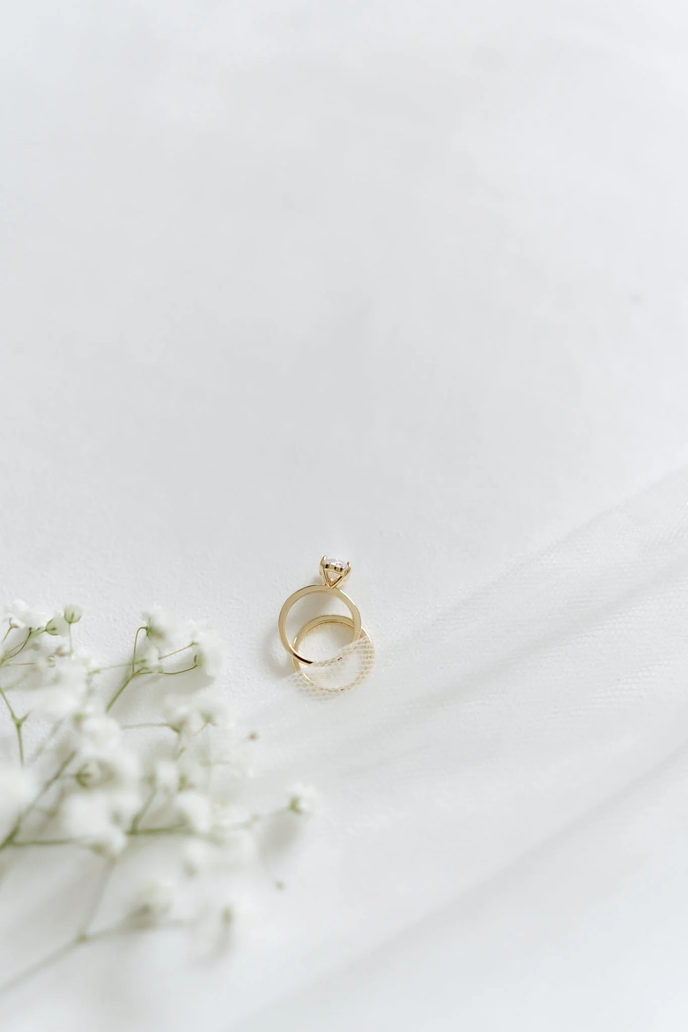 A gold ring with a diamond on top and a textured band, placed on a white surface with a sprig of baby's breath flowers nearby.