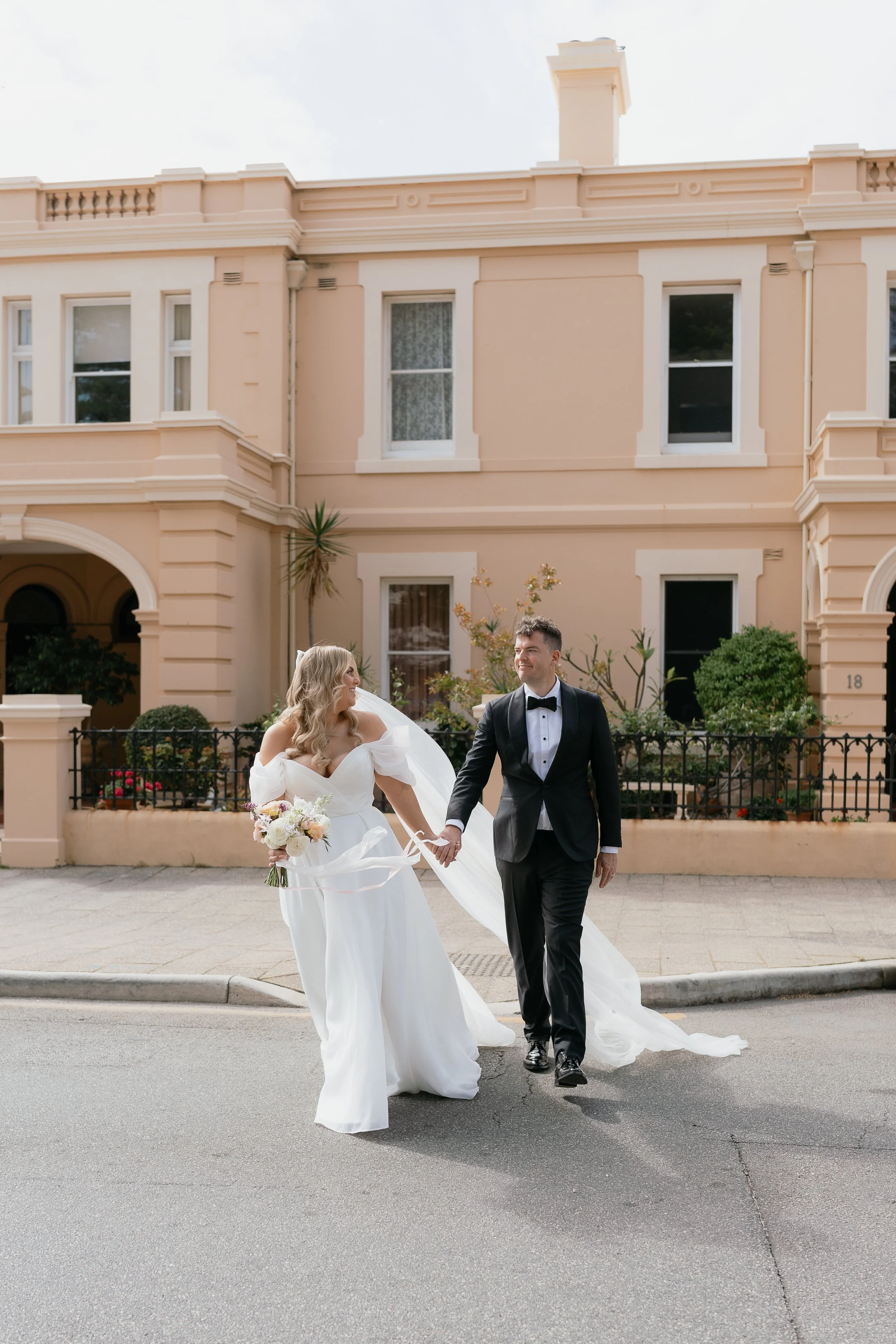 Wedding couple walking hand in hand on street, woman in white gown with bouquet, man in black tuxedo, pink historic building in background.