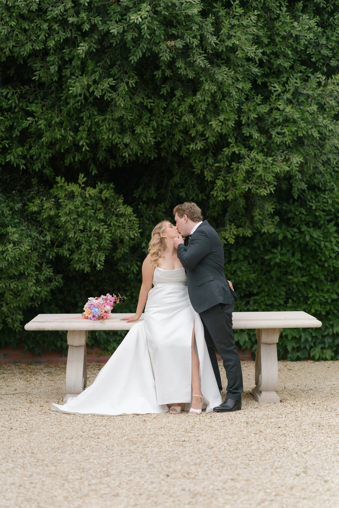 A bride and groom sharing a kiss outdoors, with a green leafy backdrop. The bride wears a white wedding gown and heels, while the groom wears a black suit. A bouquet of colorful flowers rests on the bench beside them.