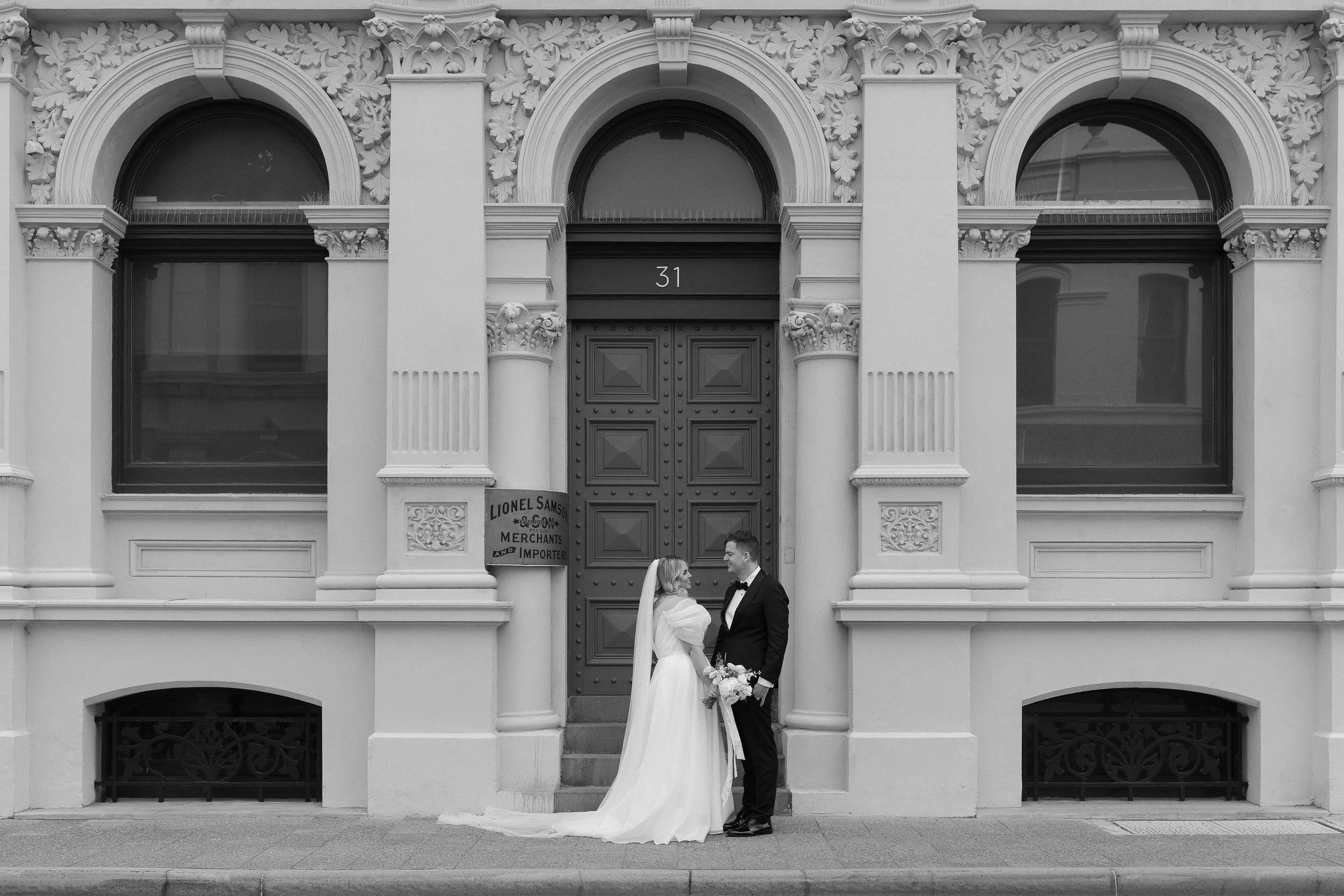 Black and white photo of a bride and groom standing in front of a historic building with ornate architectural details and a large central door. The bride is in a white wedding dress holding a bouquet, and the groom is in a black tuxedo.