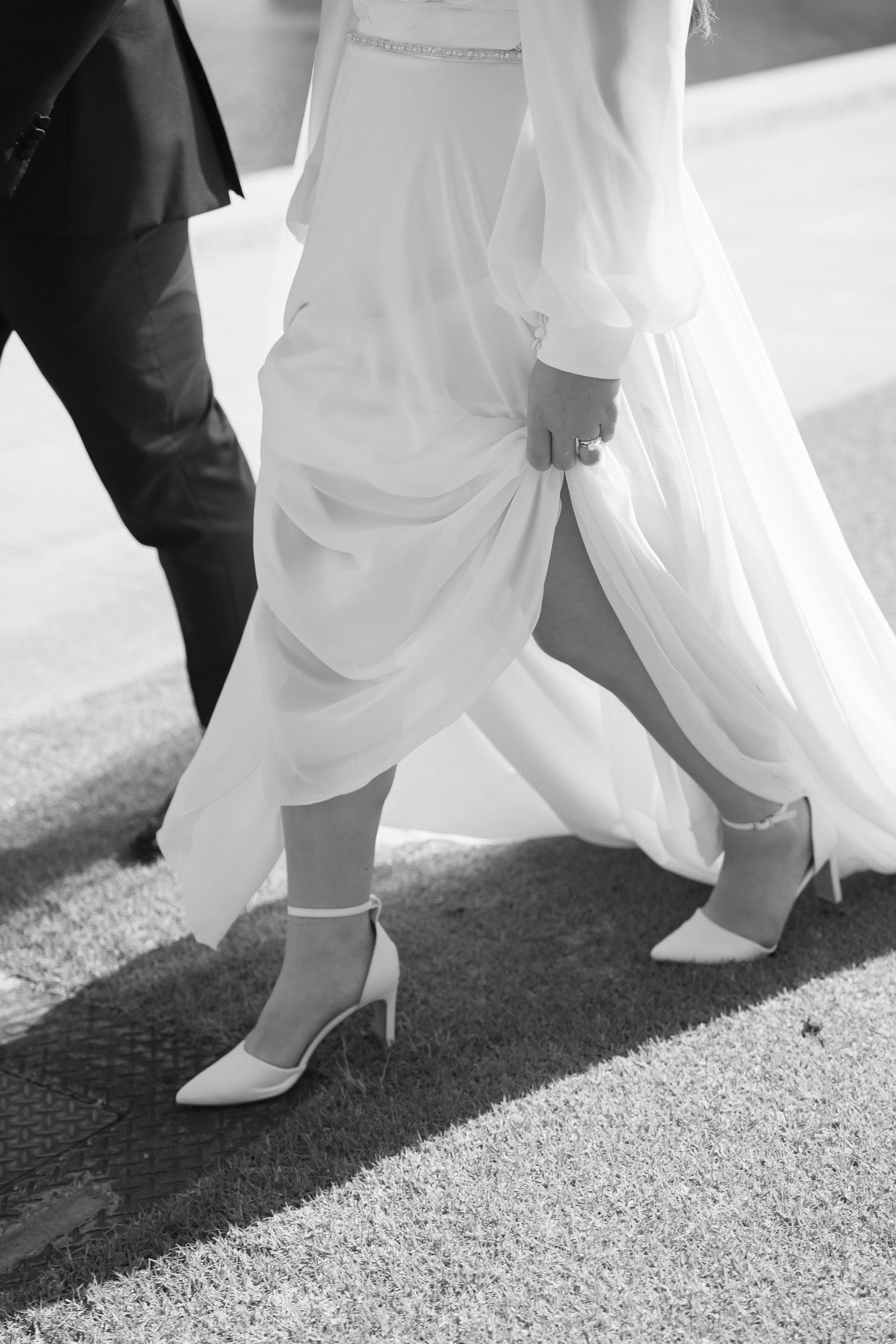Close-up of a bride lifting her wedding dress to show her leg, wearing white high heels, with a groom in a suit in the background.