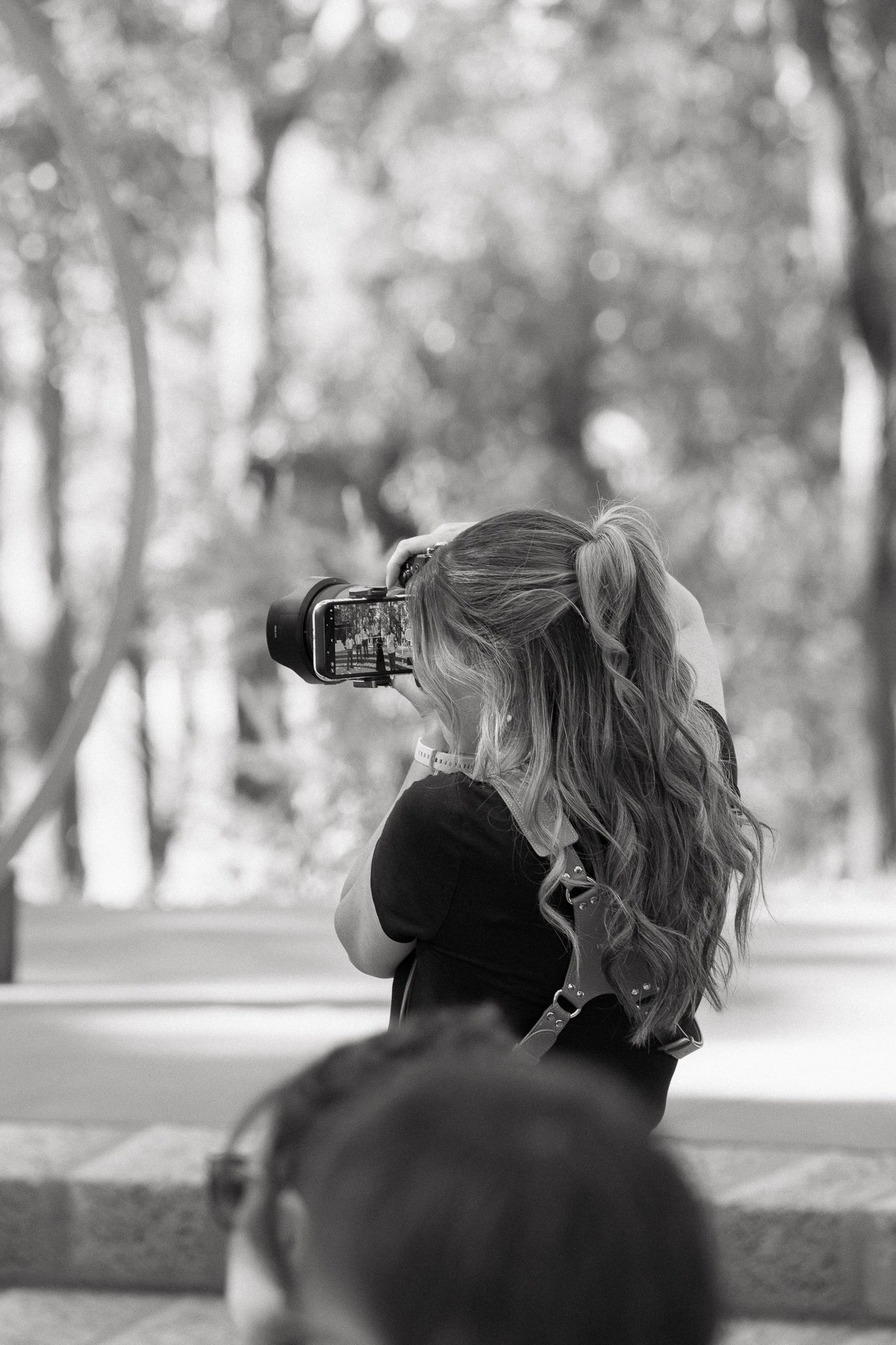 A girl with long wavy hair taking a photo with a camera outdoors, black and white image.