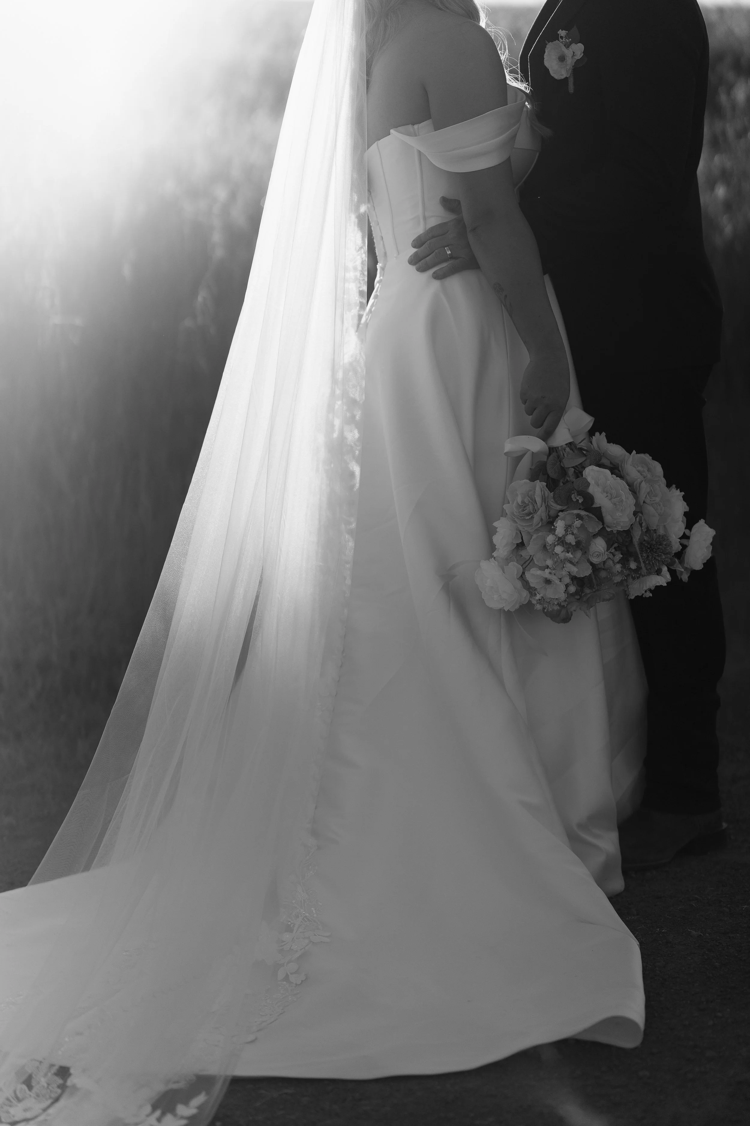 A bride and groom standing close together at sunset, the bride in a white wedding gown holding a bouquet, and the groom in a dark suit with a boutonniere, outdoors in a field.