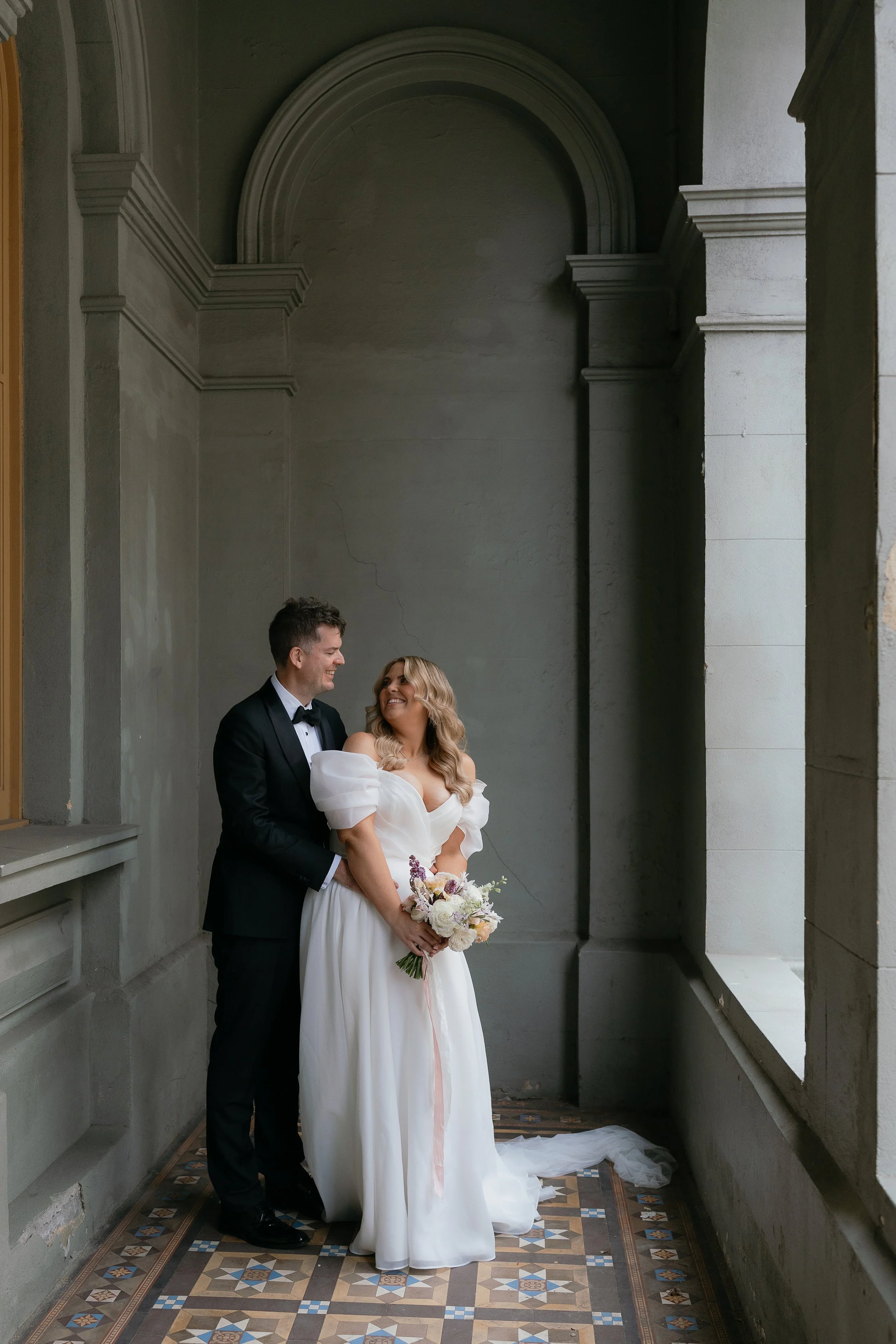 A bride and groom stand together on a patterned tile floor in a large, historic hall with high ceilings and archways. The bride wears a white wedding dress with puffed sleeves and holds a bouquet of flowers. The groom is dressed in a black tuxedo wit