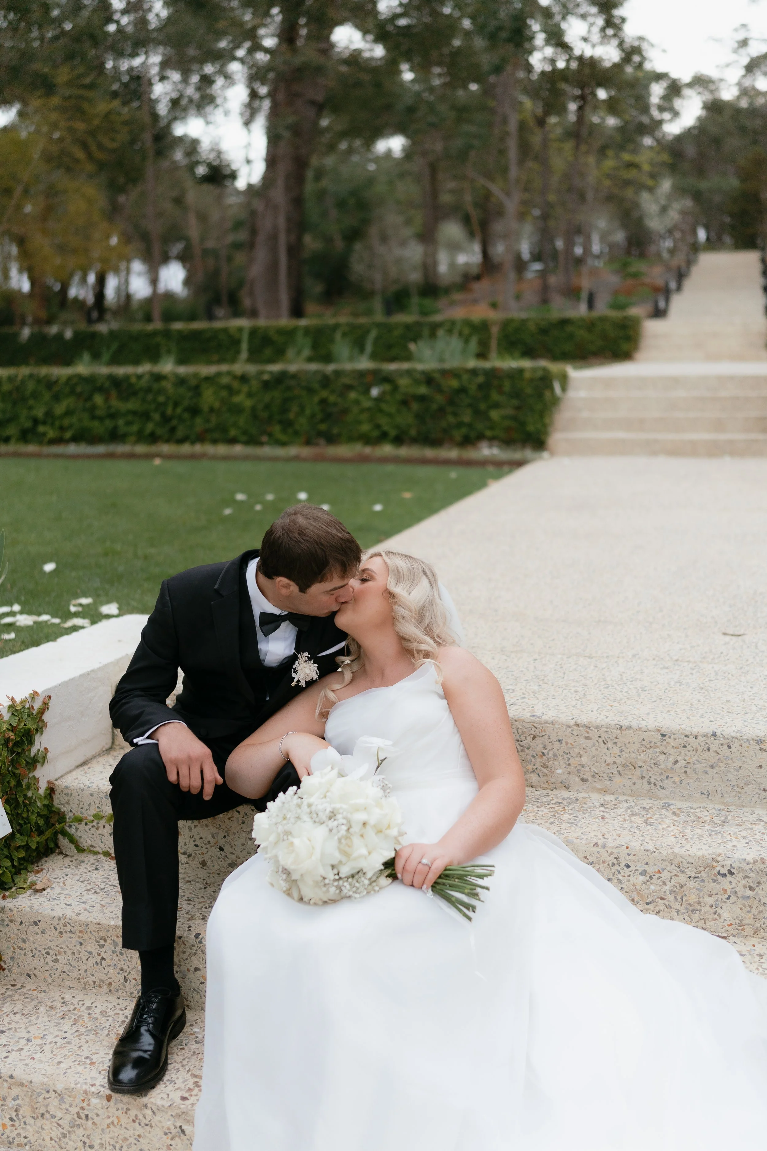 Bride and groom sitting on stairs, sharing a kiss, with the bride holding a bouquet of white flowers, outdoors in a park setting.