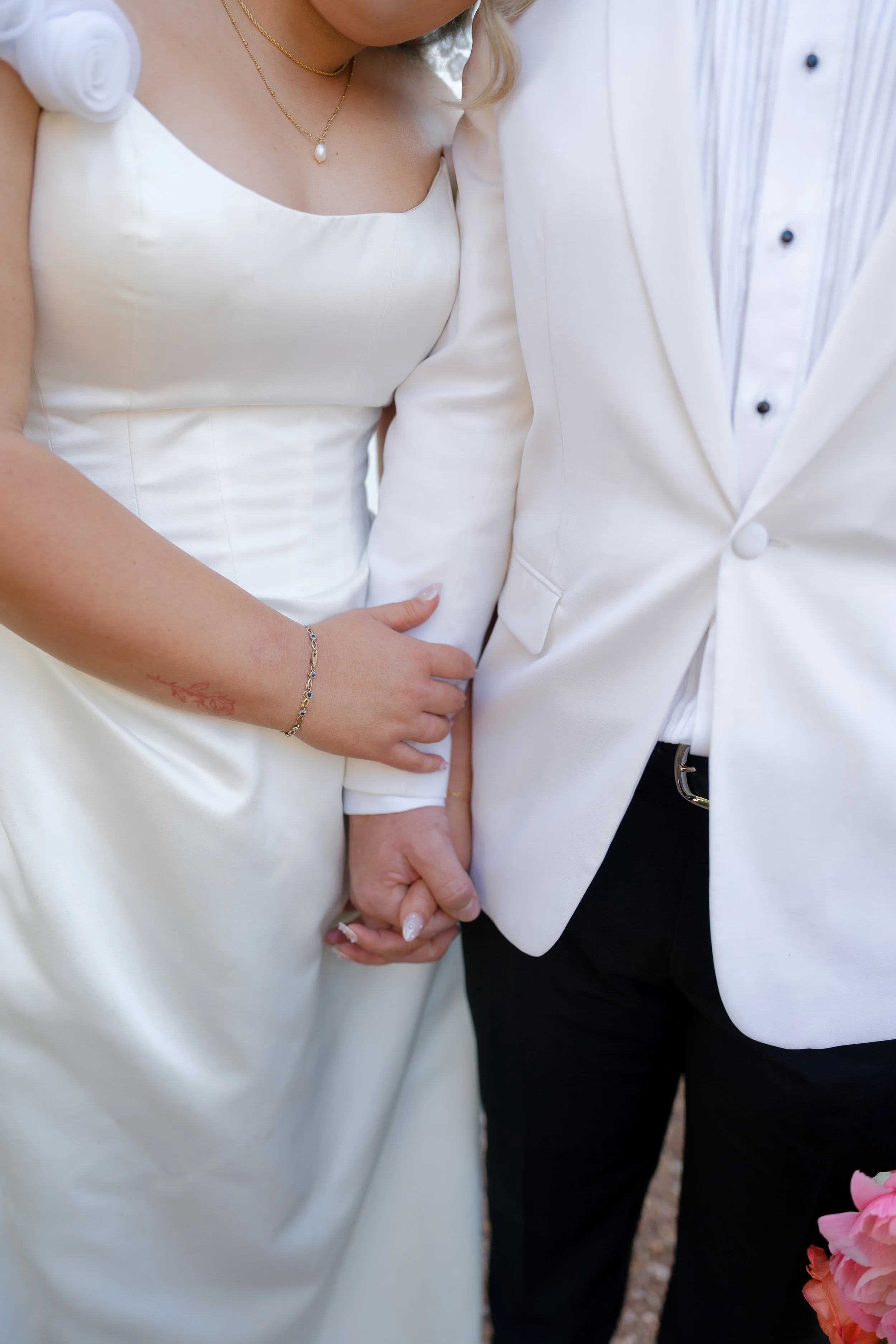 Close-up of a bride and groom holding hands, dressed in white wedding attire, with the bride's hand resting on the groom's arm.