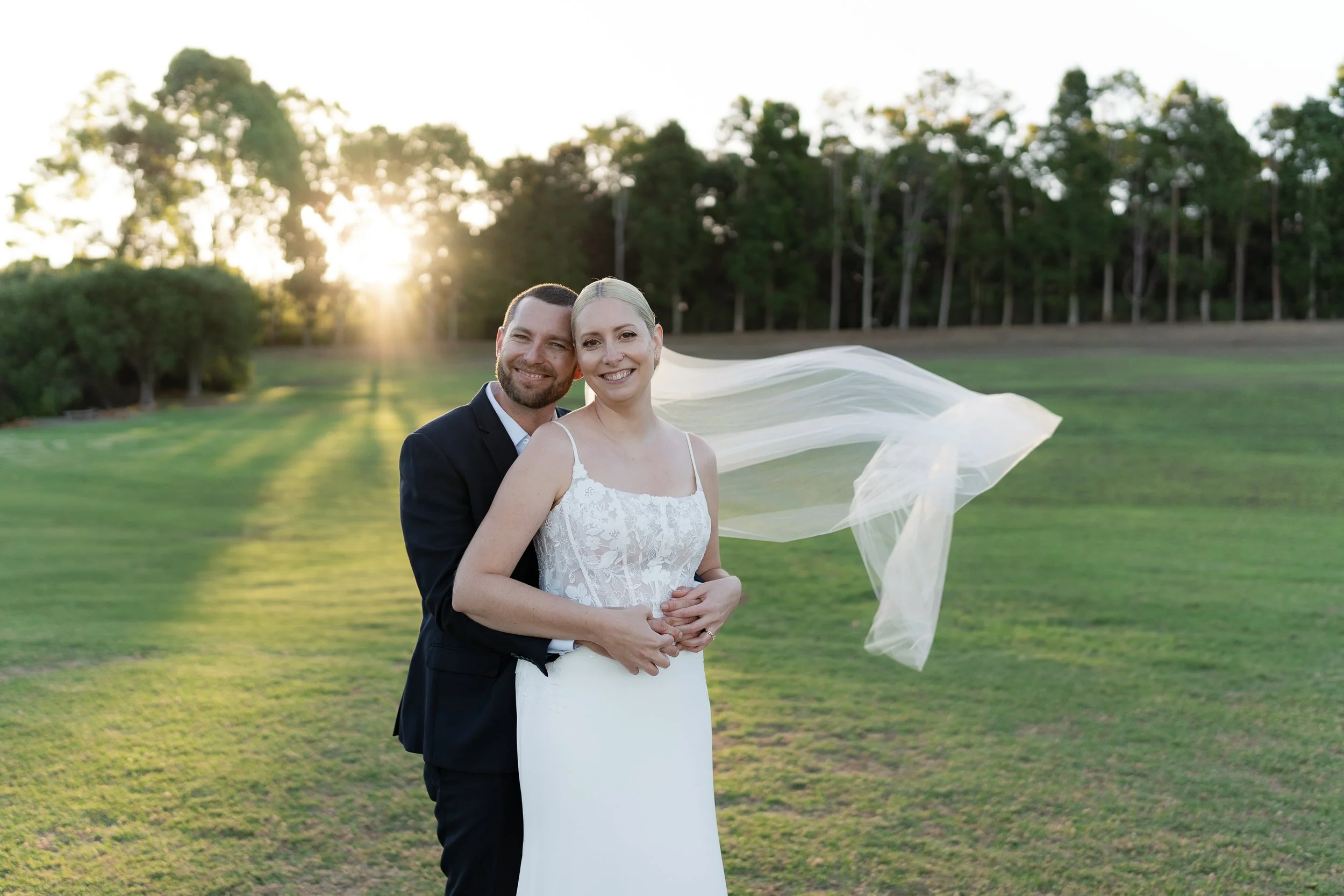 A newlywed couple stands on a grassy field during sunset, with the bride in a white dress and veil, and the groom in a black suit, smiling at the camera.