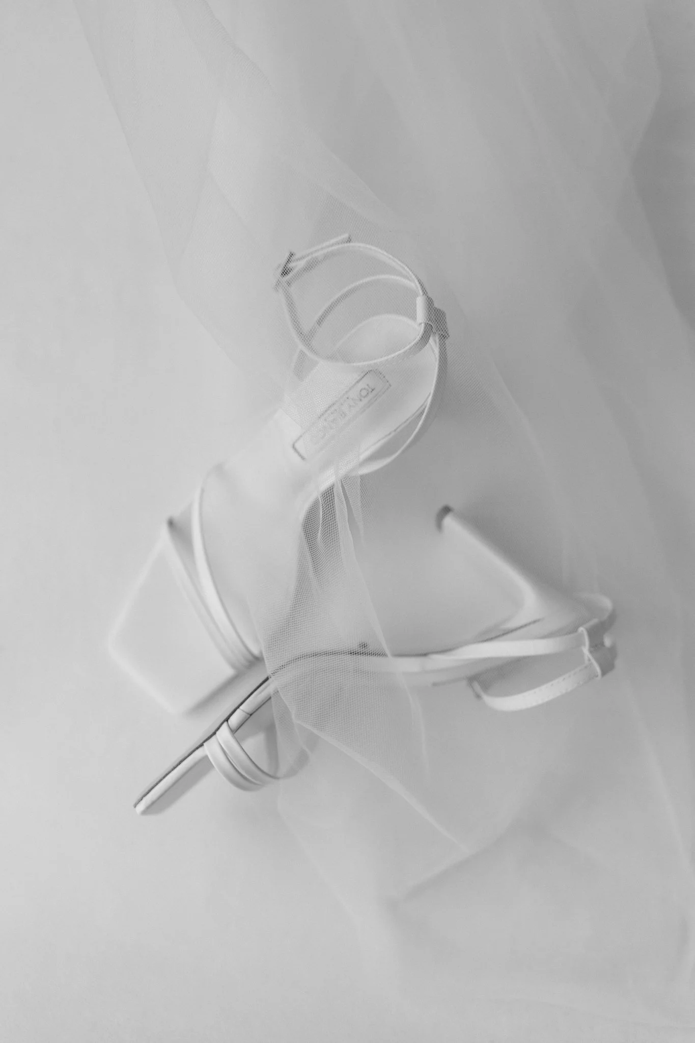 A pair of white high-heeled sandals with ankle straps, partially covered by a sheer, flowing fabric.
