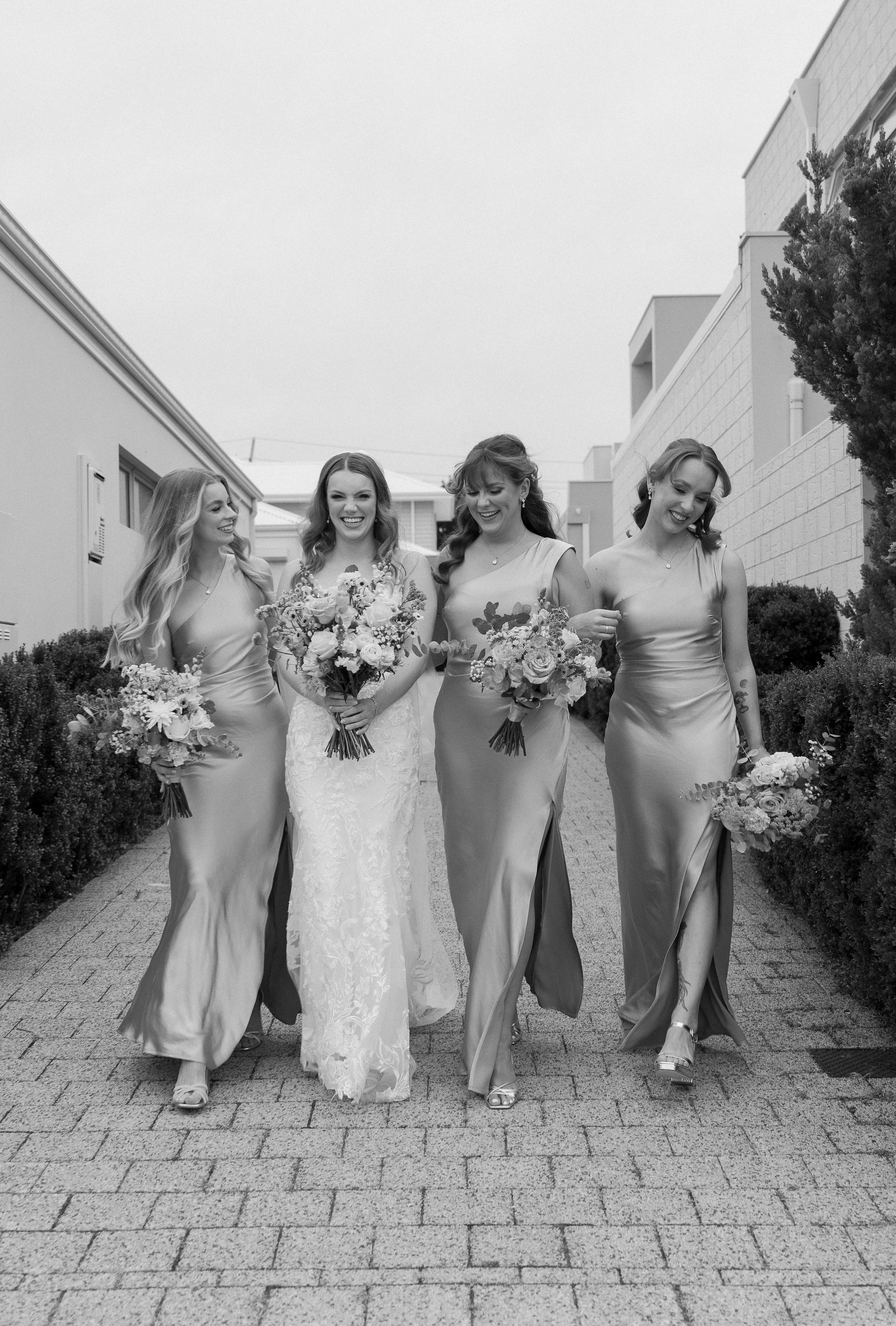 Four women in evening gowns walking outdoors, smiling, and holding bouquets of flowers.