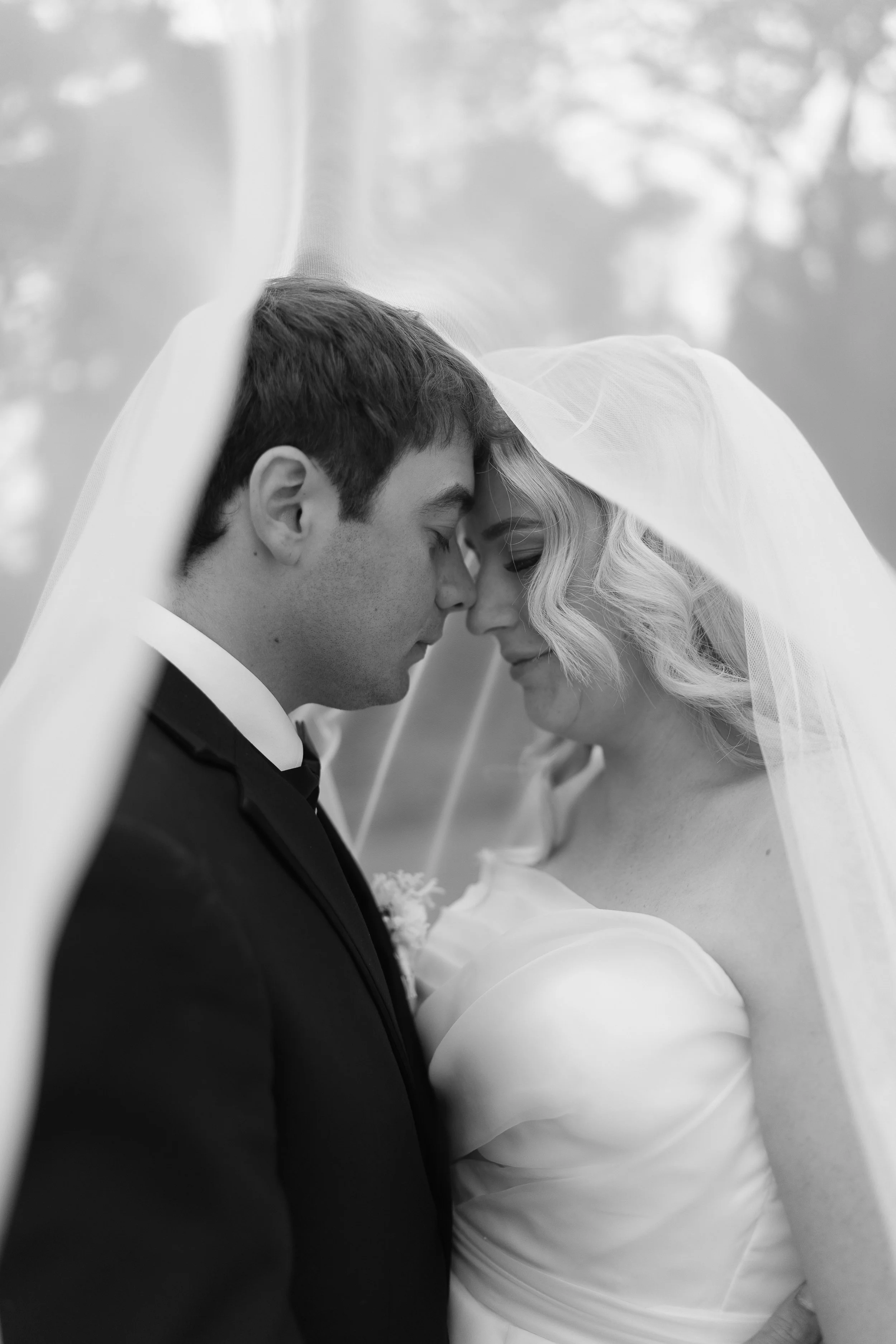 Black and white photo of a bride and groom with foreheads touching under a veil