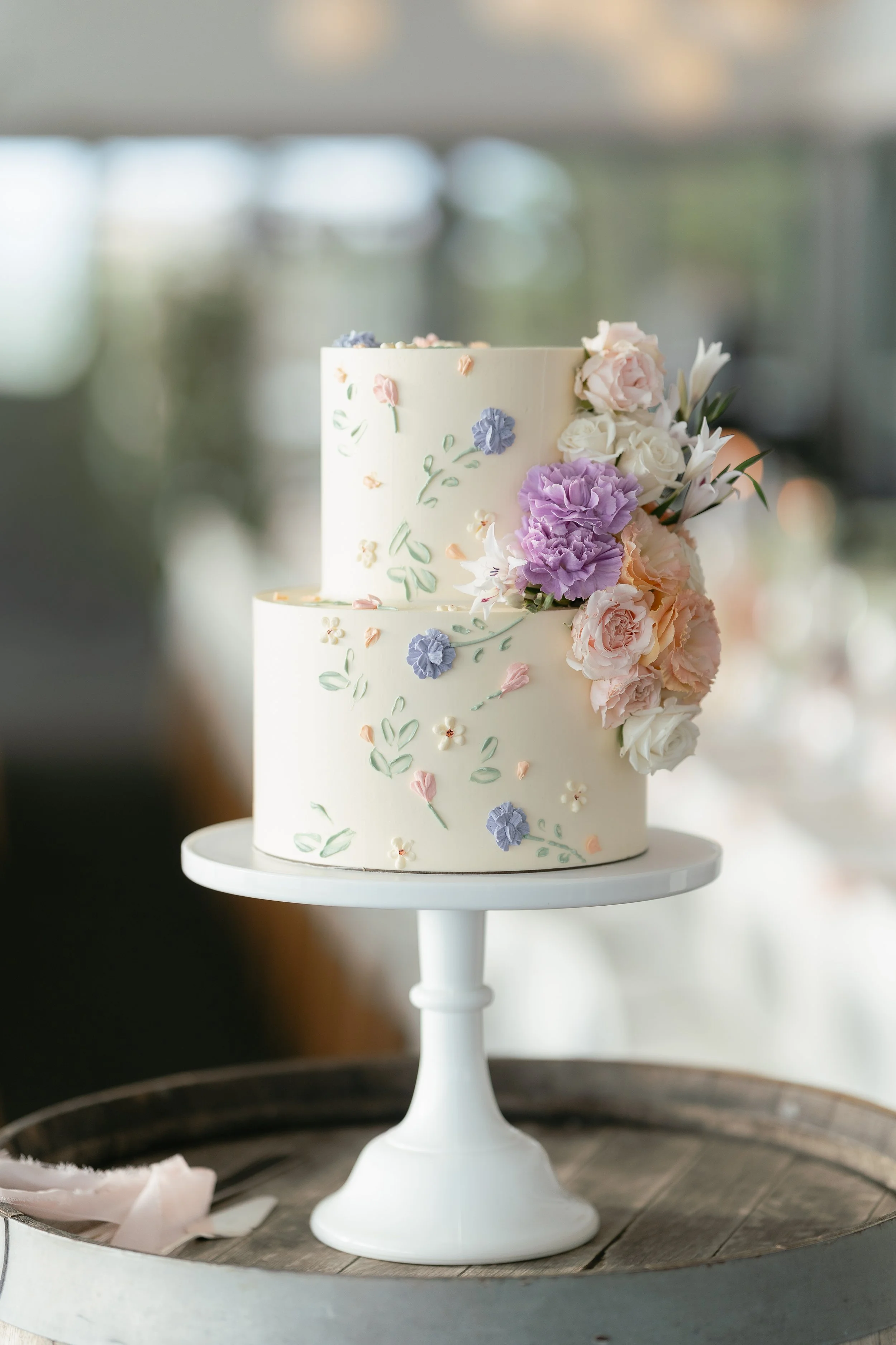 A two-tier white wedding cake decorated with pastel-colored flowers and green leaves, displayed on a white cake stand on a wooden table.