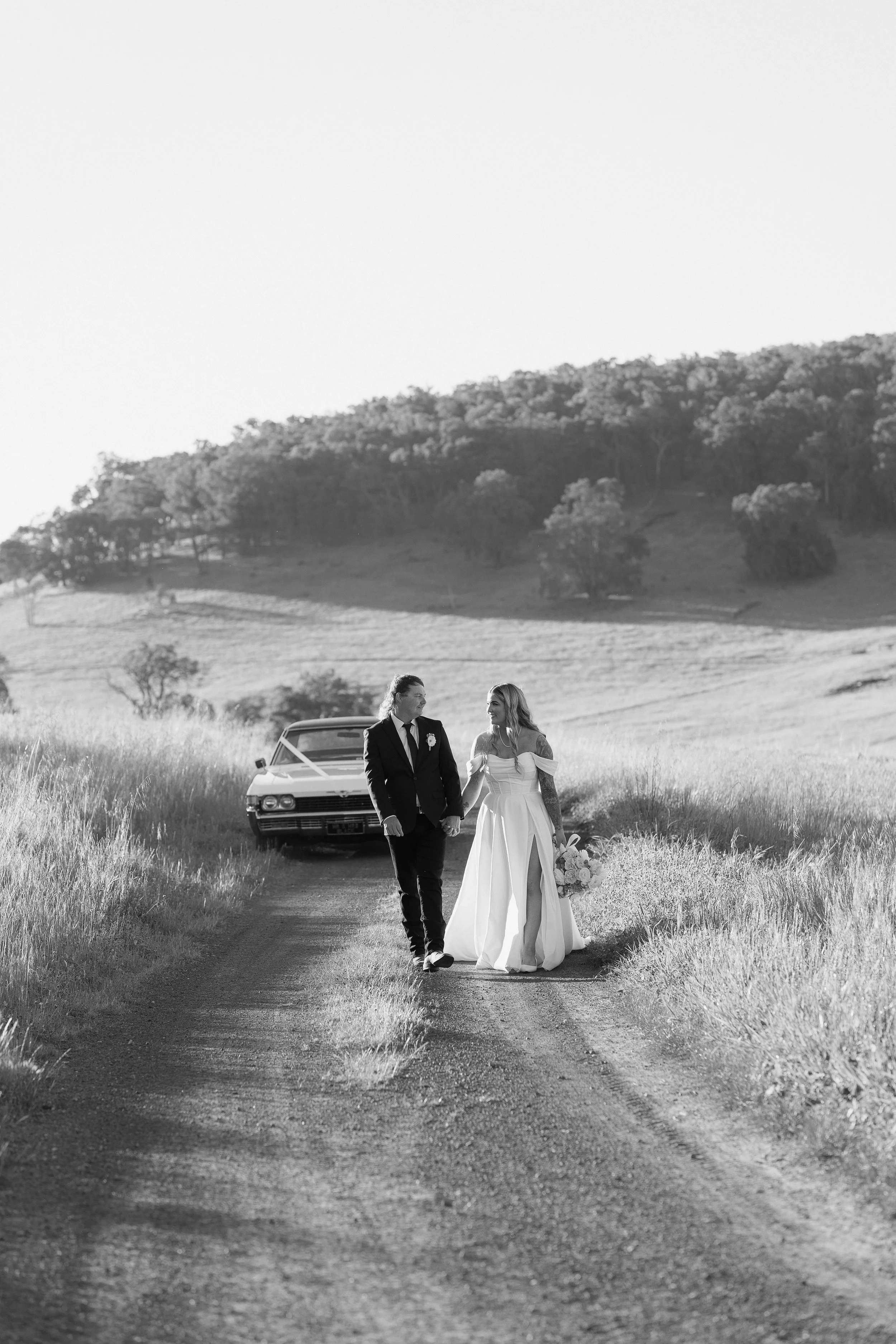 Black and white photo of a bride and groom walking hand in hand on a dirt path through a rural landscape with trees on hills in the background, wedding dress, bouquet of flowers, and wedding attire.