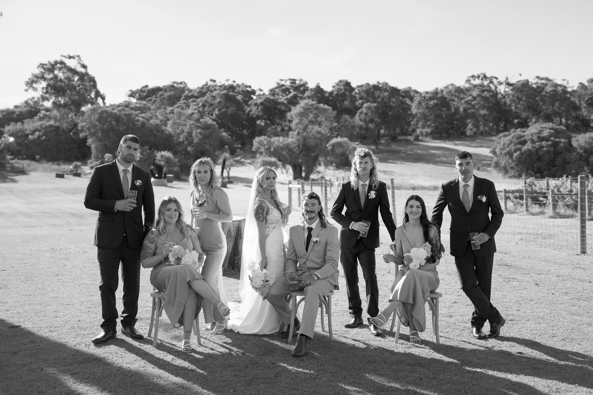Black and white photo of a group of wedding party members outdoors, including a bride in a wedding gown, groom in a suit, three women and three men, some holding drinks and flowers, with trees and open fields in the background.