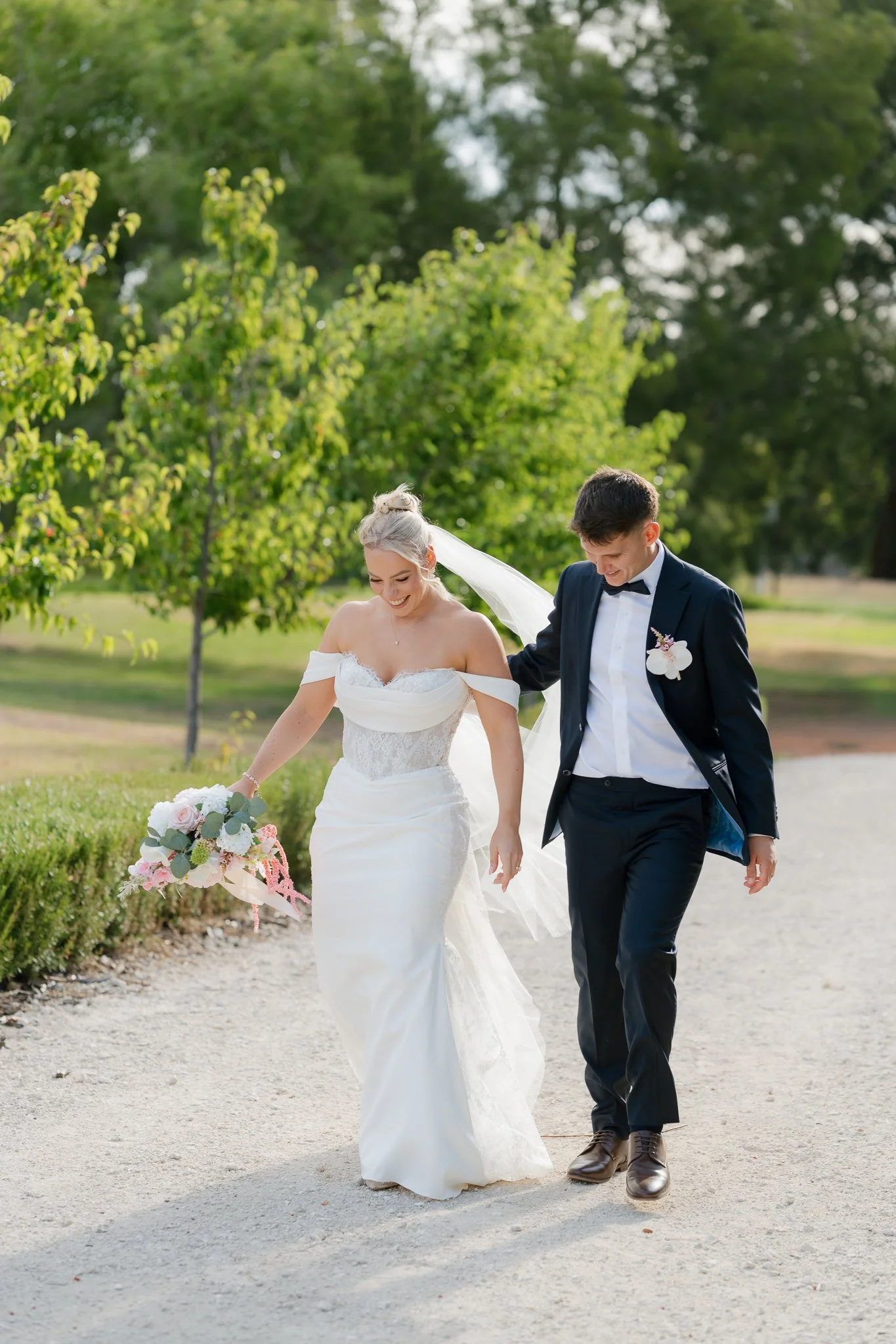 A bride and groom walking outdoors on their wedding day, smiling and holding each other's arm. The bride is in a white wedding dress holding a bouquet of pink and white flowers, and the groom is in a black tuxedo with a bow tie and boutonniere. There