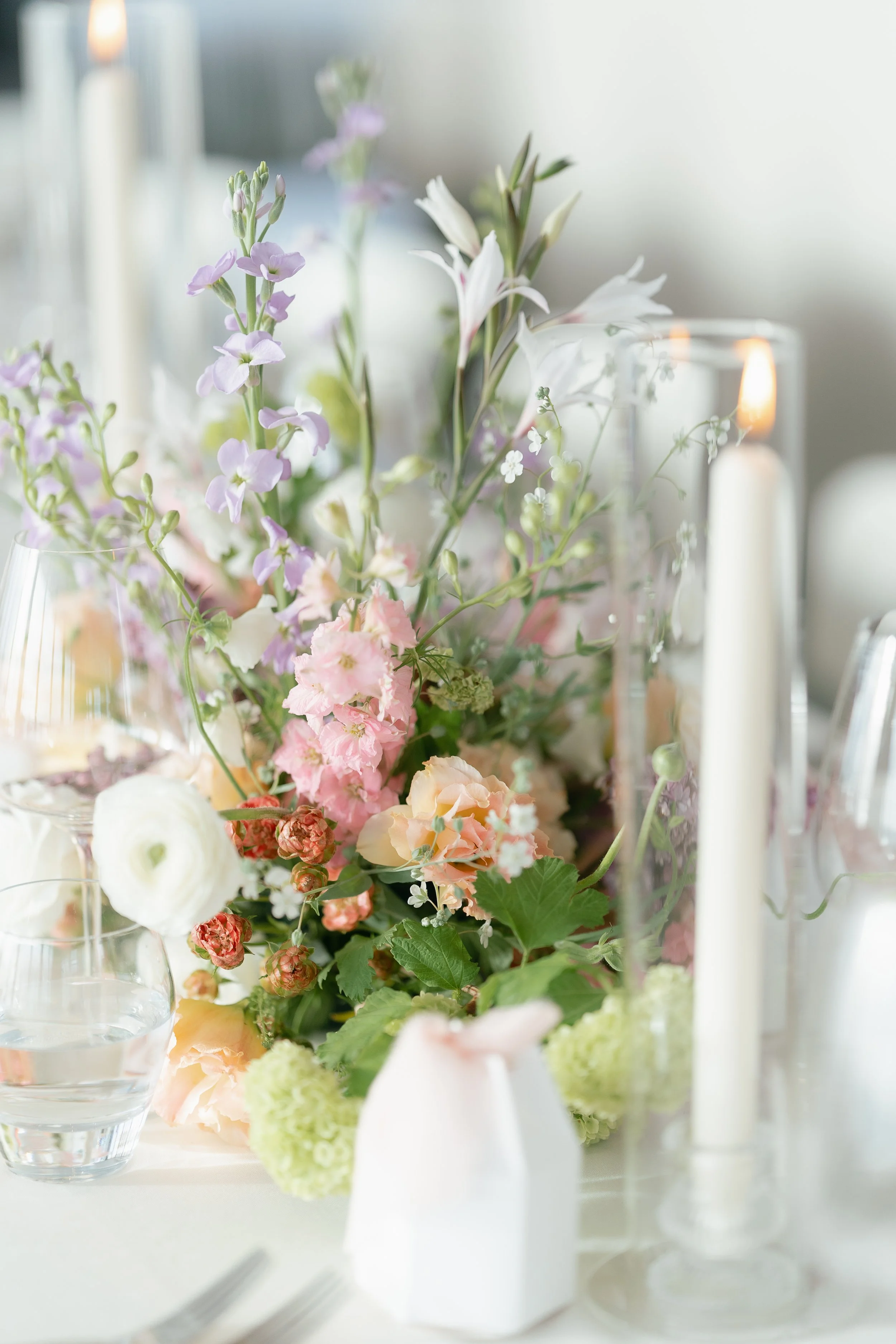 A floral centerpiece with pink, white, and lavender flowers on a table, accompanied by candles and glassware.