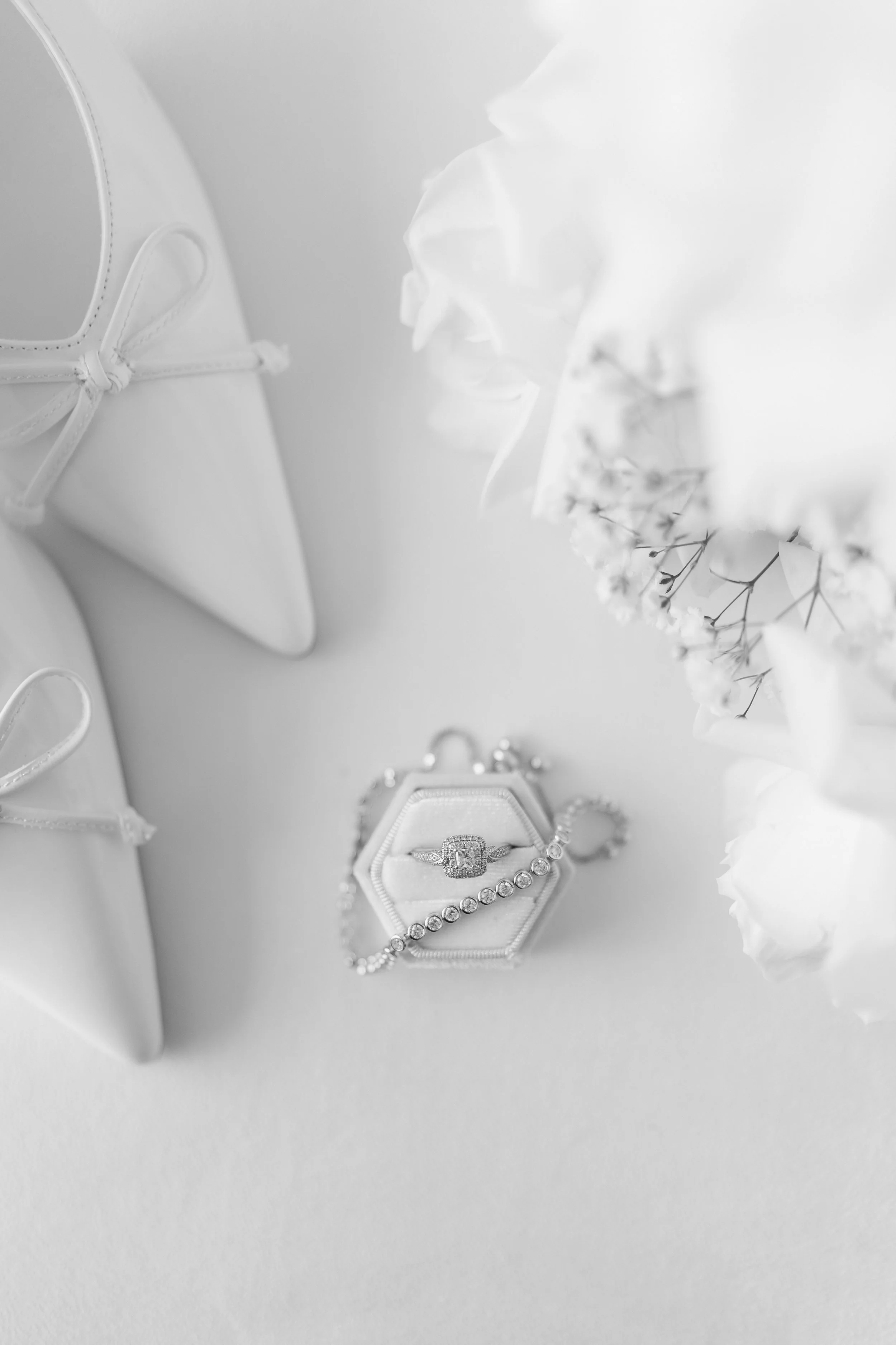 Black and white photo of a jewelry ring in a hexagonal box, with a pearl bracelet, white shoes, and flowers nearby.