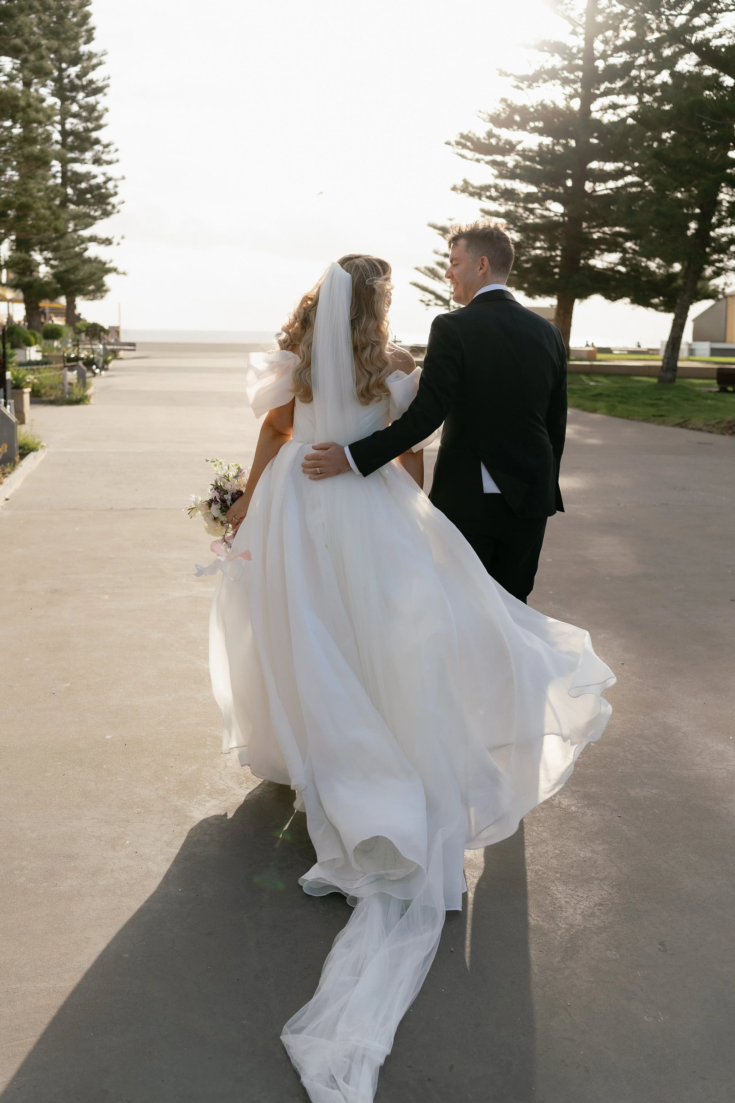 A bride and groom walking outdoors on a sunny day, with the bride in a white wedding gown holding a bouquet, and the groom in a black tuxedo.