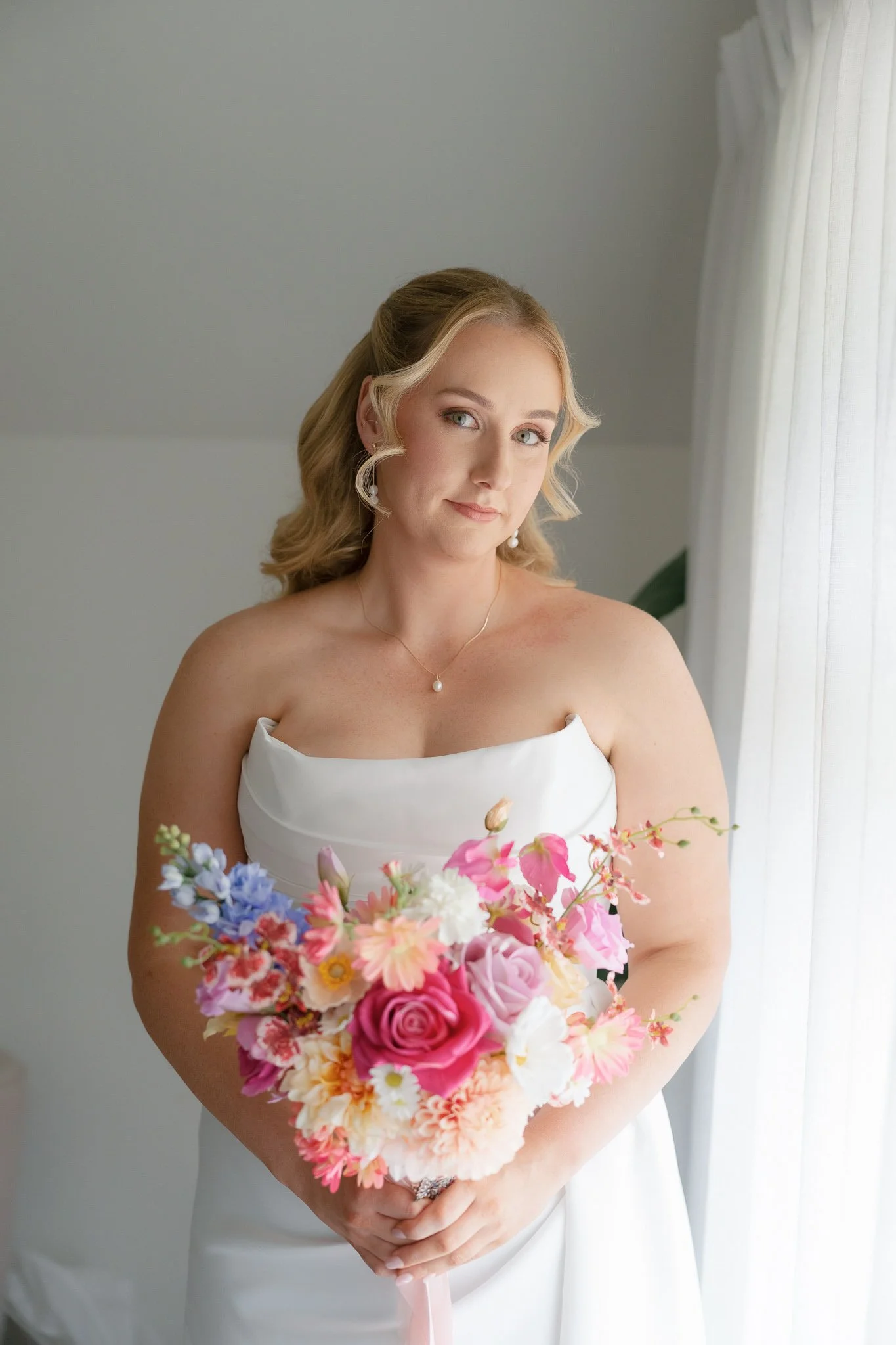 A bride with blonde hair styled in loose waves, wearing a strapless white wedding dress, holding a colorful bouquet of pink, peach, white, and purple flowers, standing near a window with white curtains.