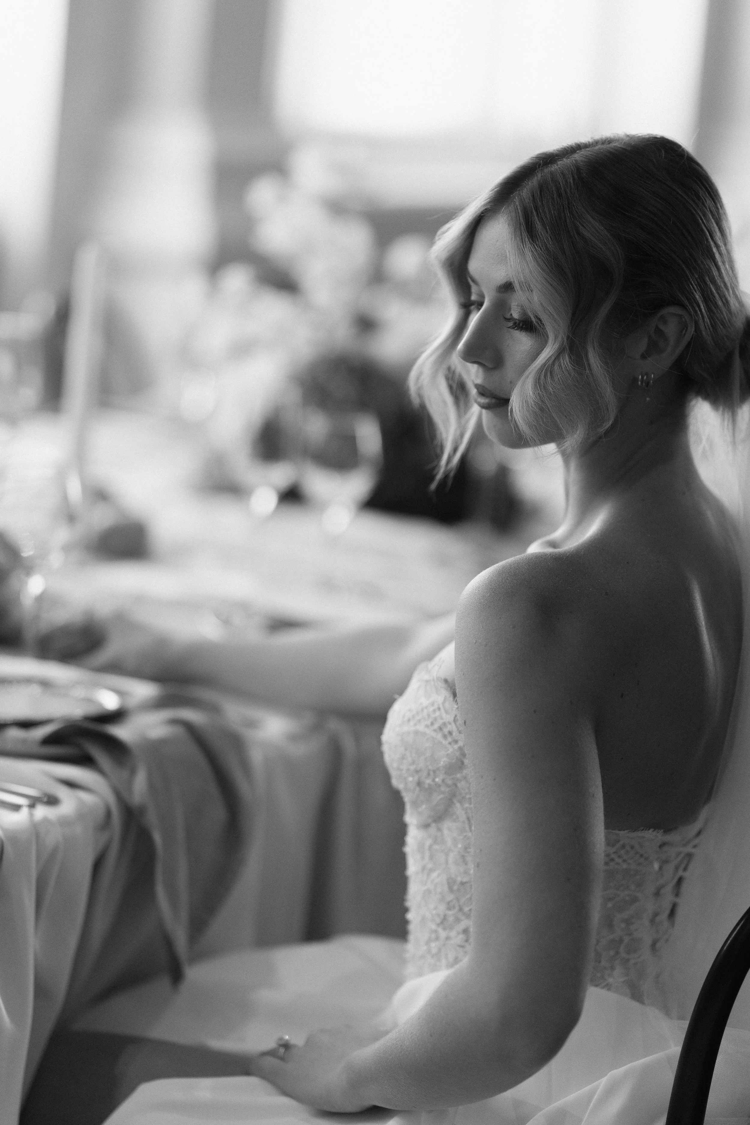 Black and white photo of a woman with styled hair sitting at a dining table, dressed in lace, with a contemplative expression.