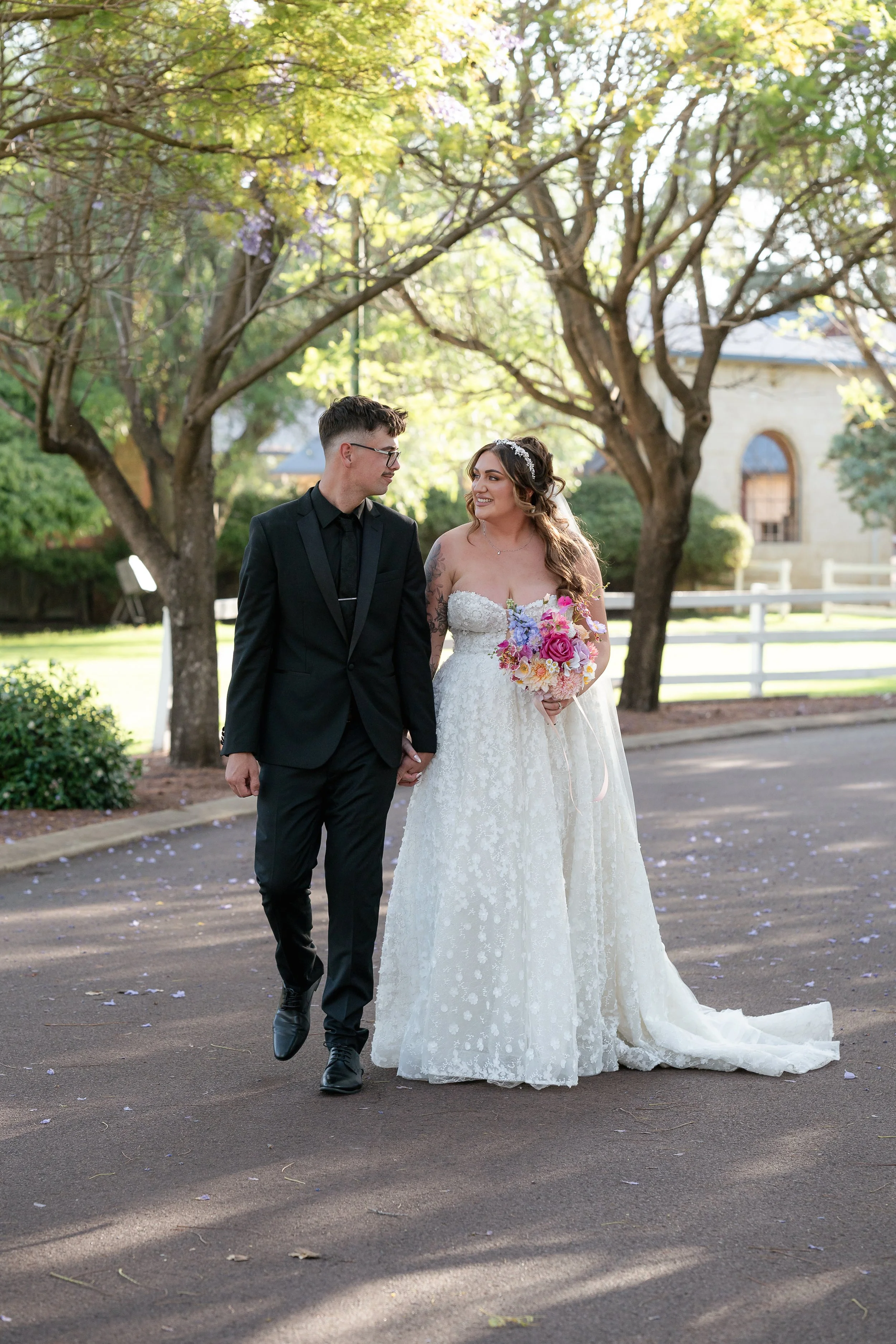 A newlywed couple walking outdoors on a sunny day with trees and a building in the background. The bride is wearing a white lace strapless wedding dress and holding a colorful bouquet, while the groom is dressed in a black suit and glasses, holding h