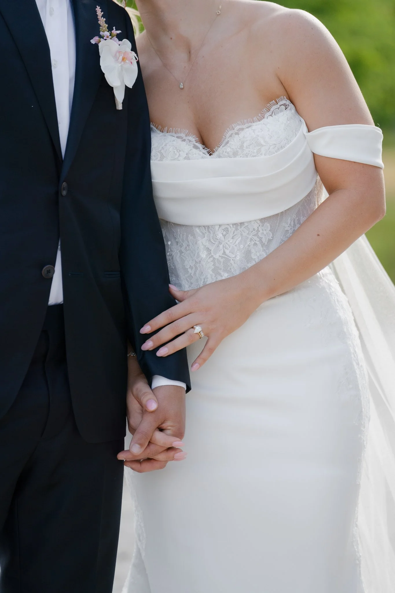 Close-up of a bride and groom holding hands. The bride is wearing a white wedding dress with lace details and an off-the-shoulder design, and a diamond ring. The groom is dressed in a black tuxedo with a pink orchid boutonniere. The couple is outdoor