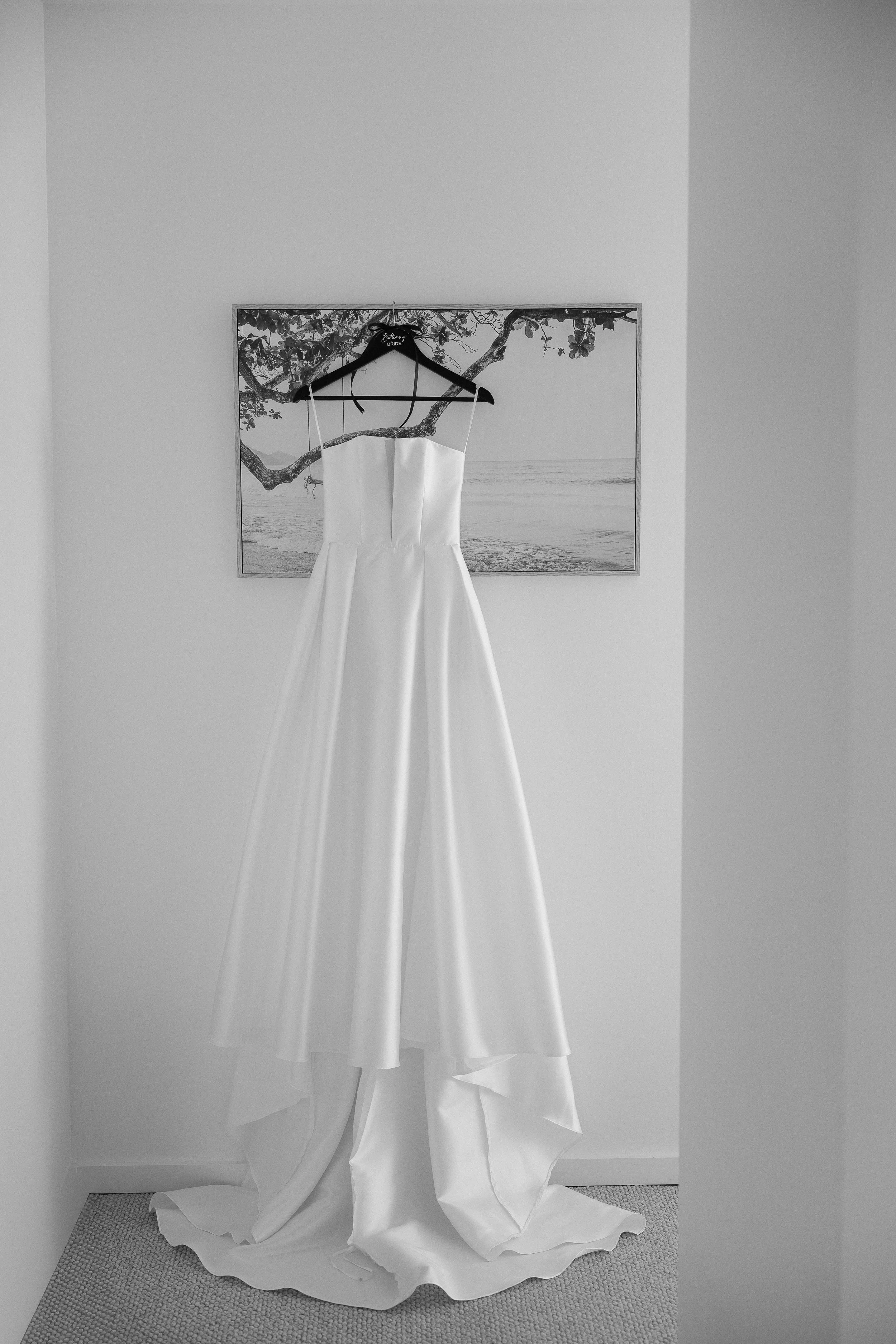 A wedding dress hanging on a black hanger on a wall, with a framed beach scene in the background.