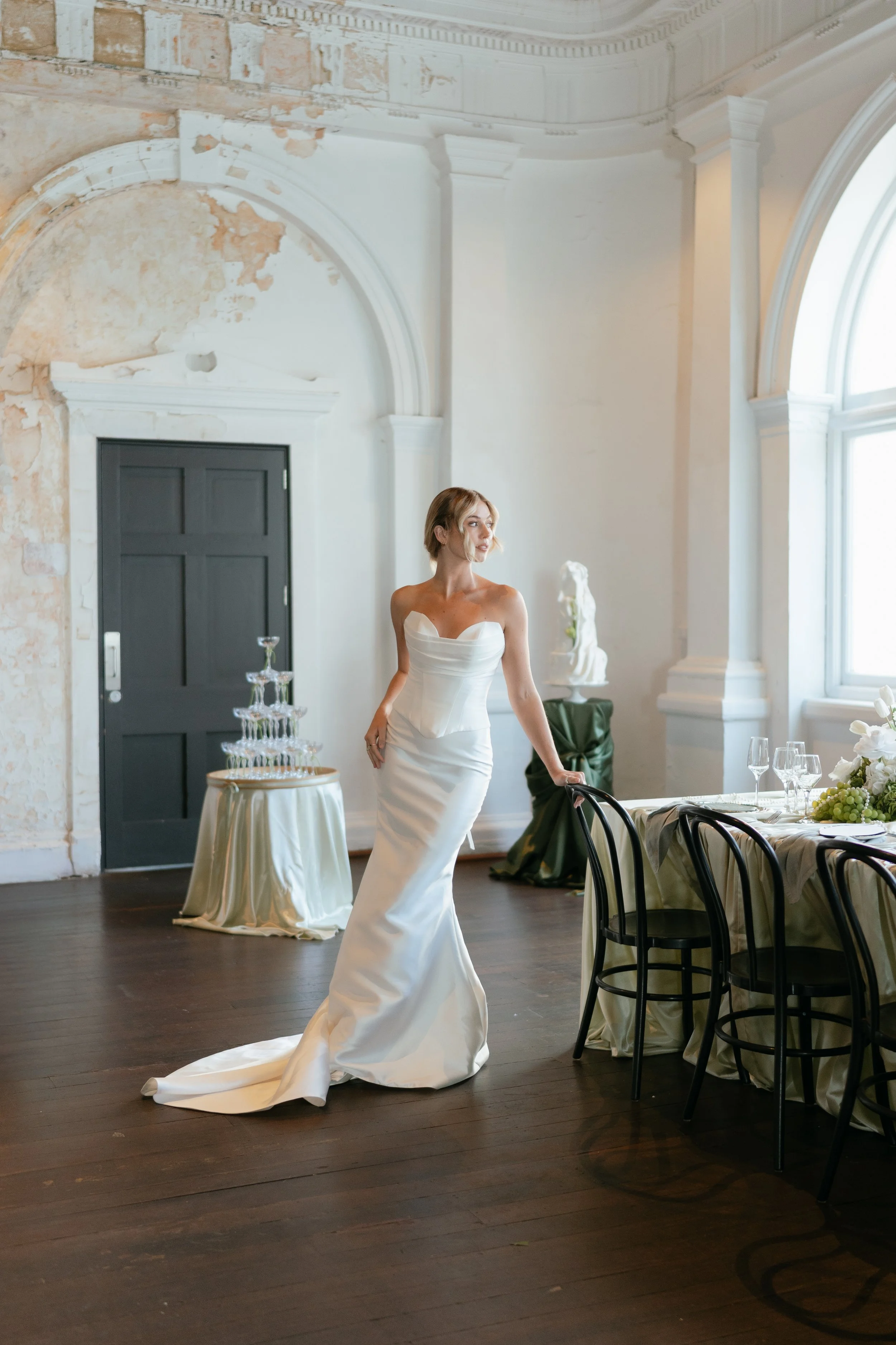 A woman in a white wedding gown standing beside a table with champagne glasses in a decorated event space with large windows and antique decor.