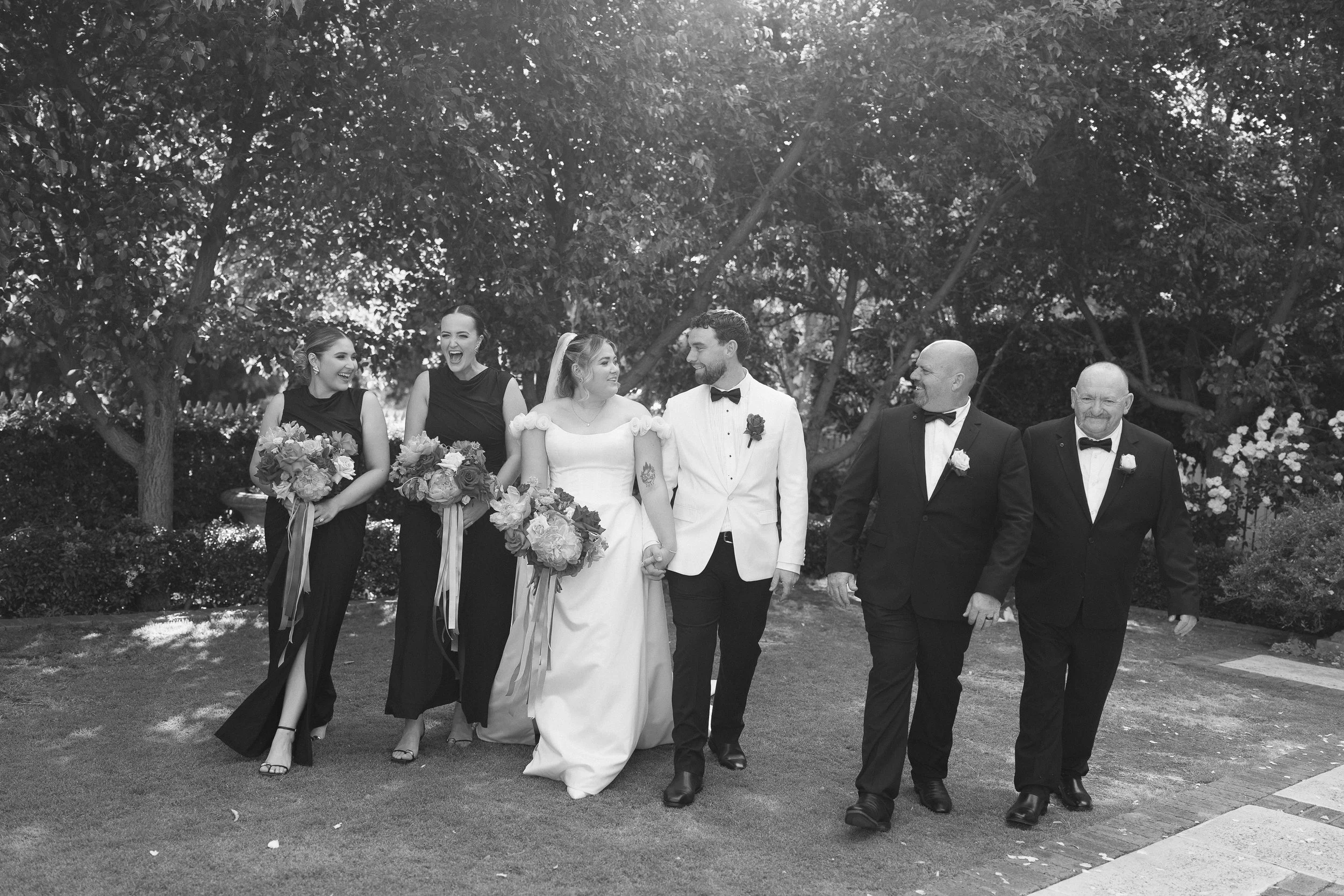 Black and white photo of a wedding party walking outdoors, including a bride in a white gown holding a bouquet, a groom in a white tuxedo with a boutonniere, two female bridesmaids in black dresses holding bouquets, and two groomsmen in black tuxedos