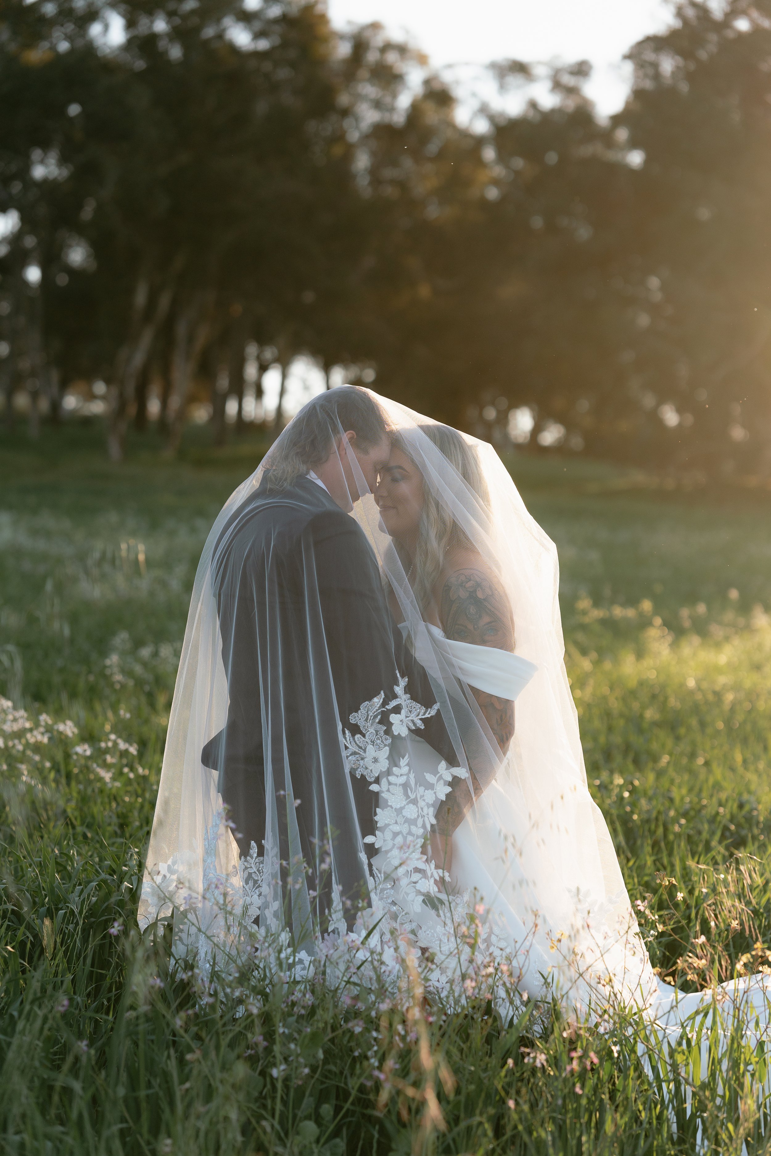 A bride and groom standing close together in a grassy field, under a veil, with trees in the background during sunset.