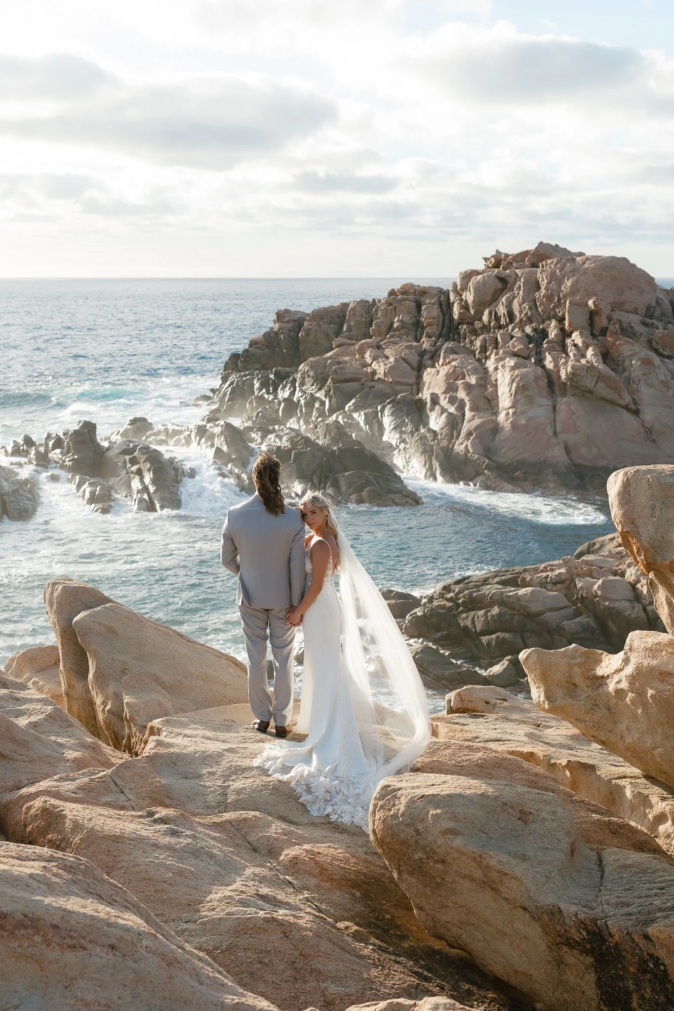 A bride and groom standing on rocks by the ocean, holding hands, with rocky cliffs and the sea in the background.