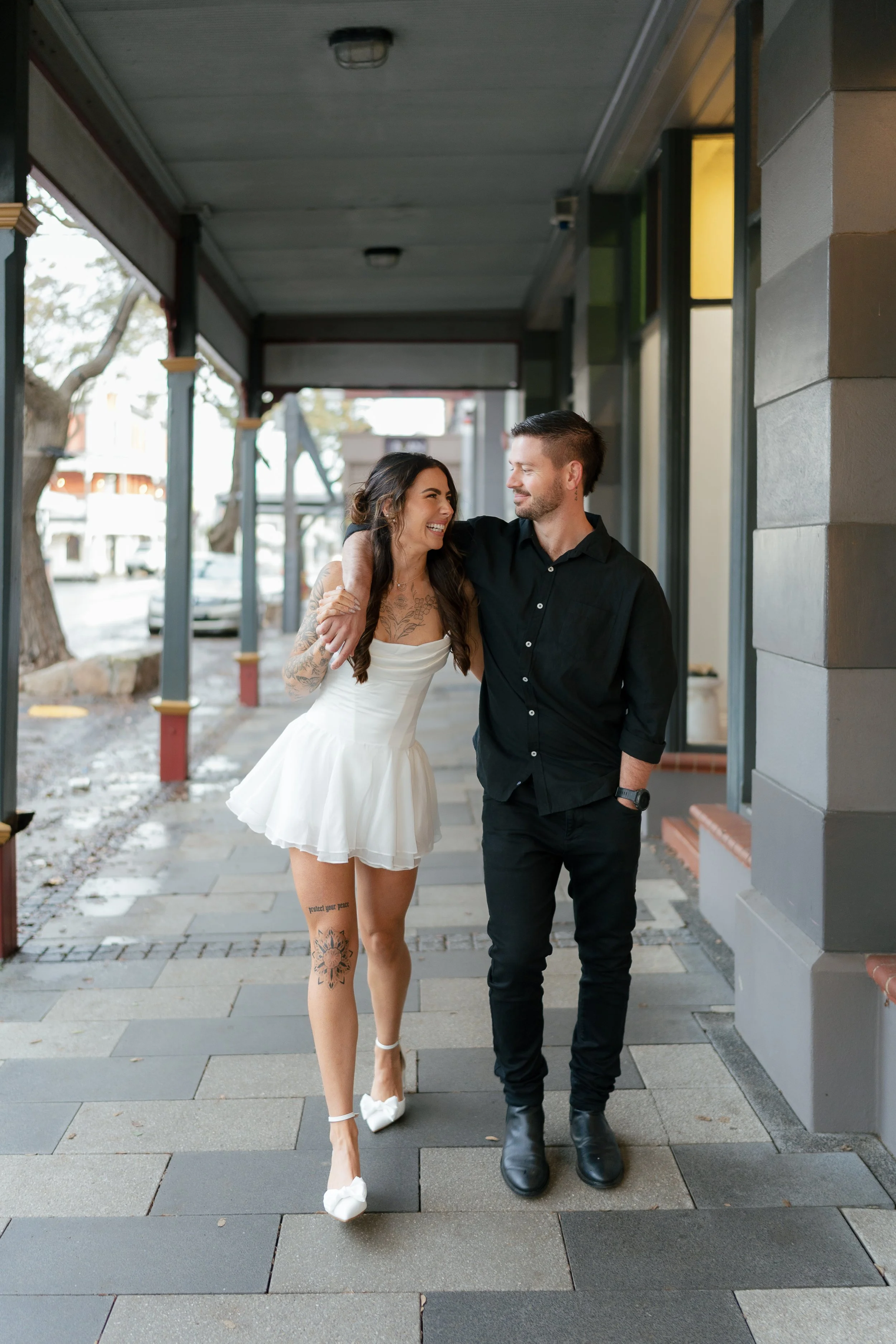 A couple walks arm-in-arm on a city sidewalk, smiling at each other. The woman is wearing a white dress and heels, while the man is dressed in black and wearing boots. They appear happy and close.