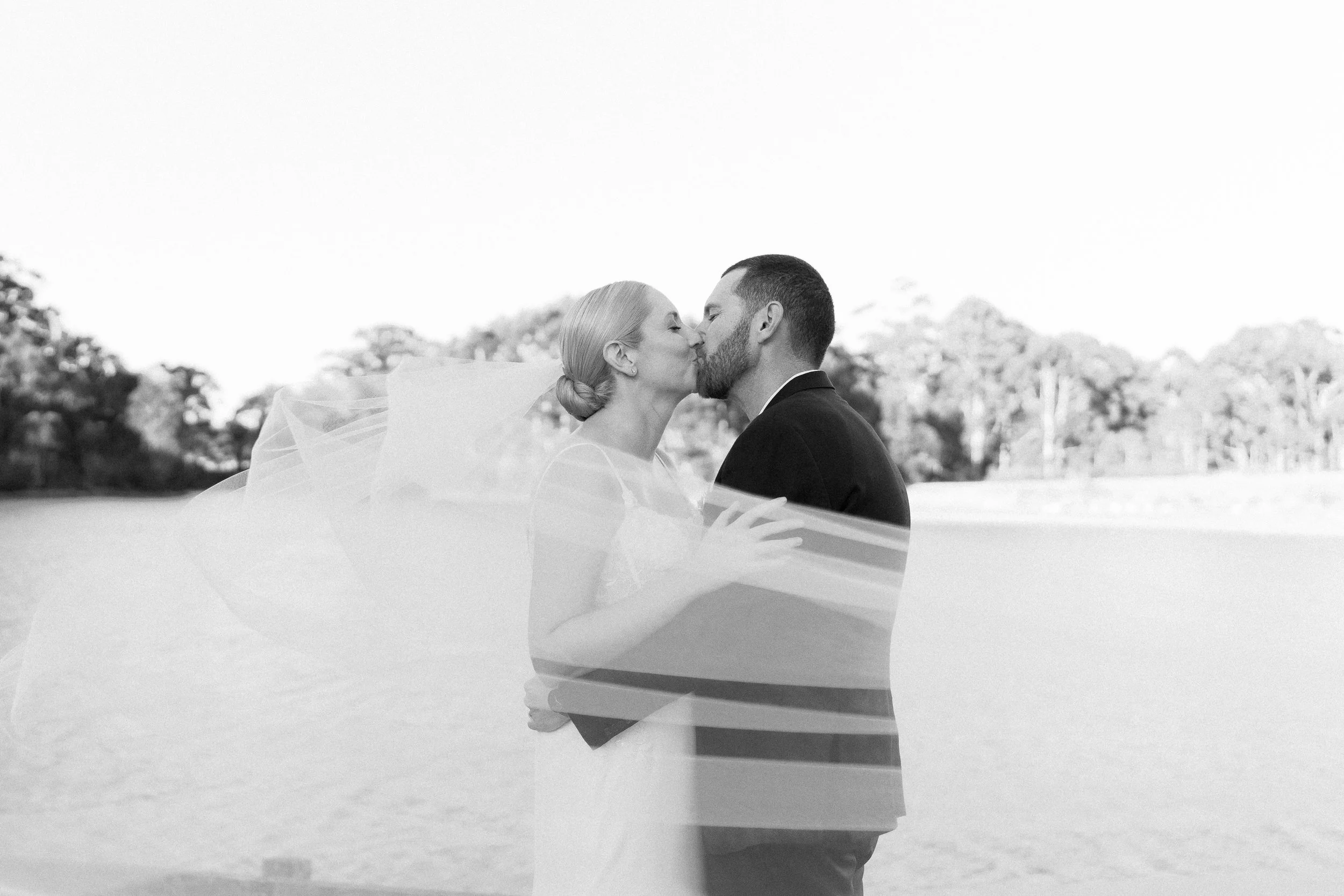 A black and white photo of a bride and groom sharing a kiss outdoors, with trees in the background.