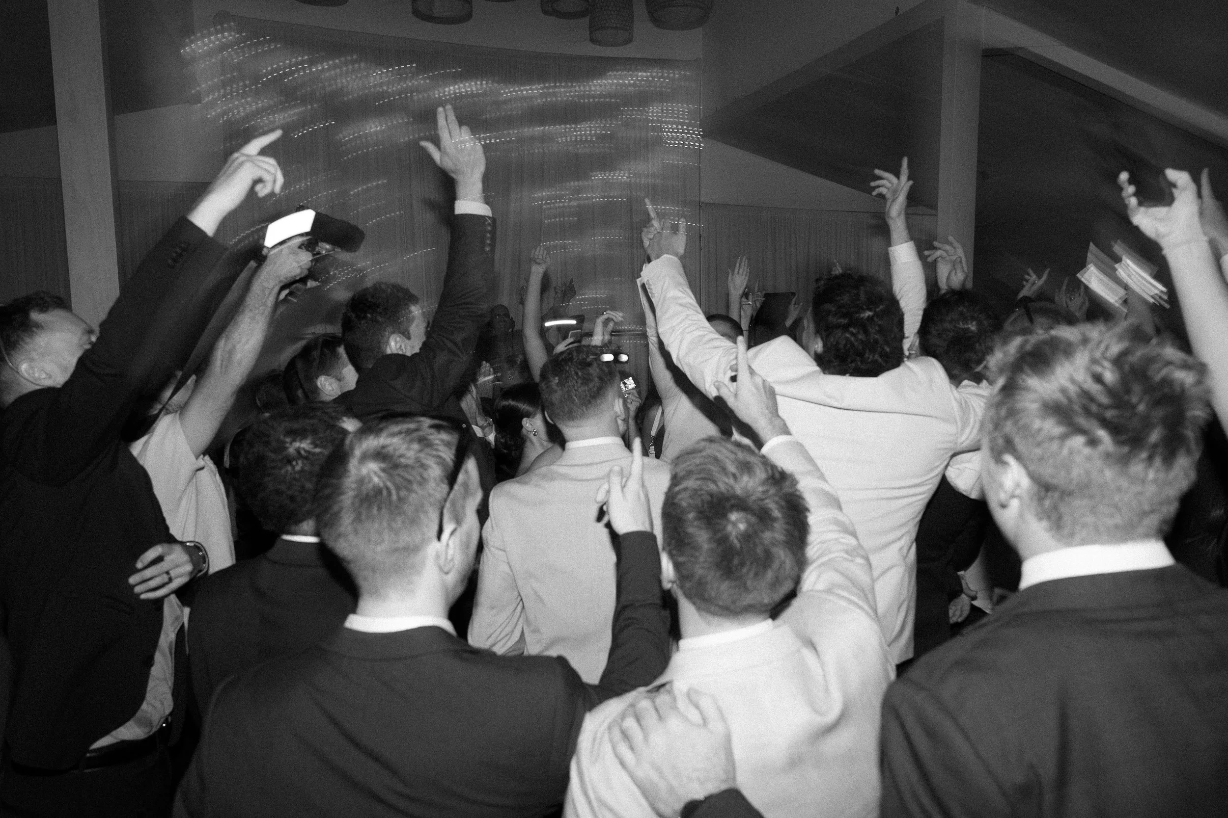 A crowd of people at a celebration or party, many with their hands raised, some taking photos, in a lively indoor setting.