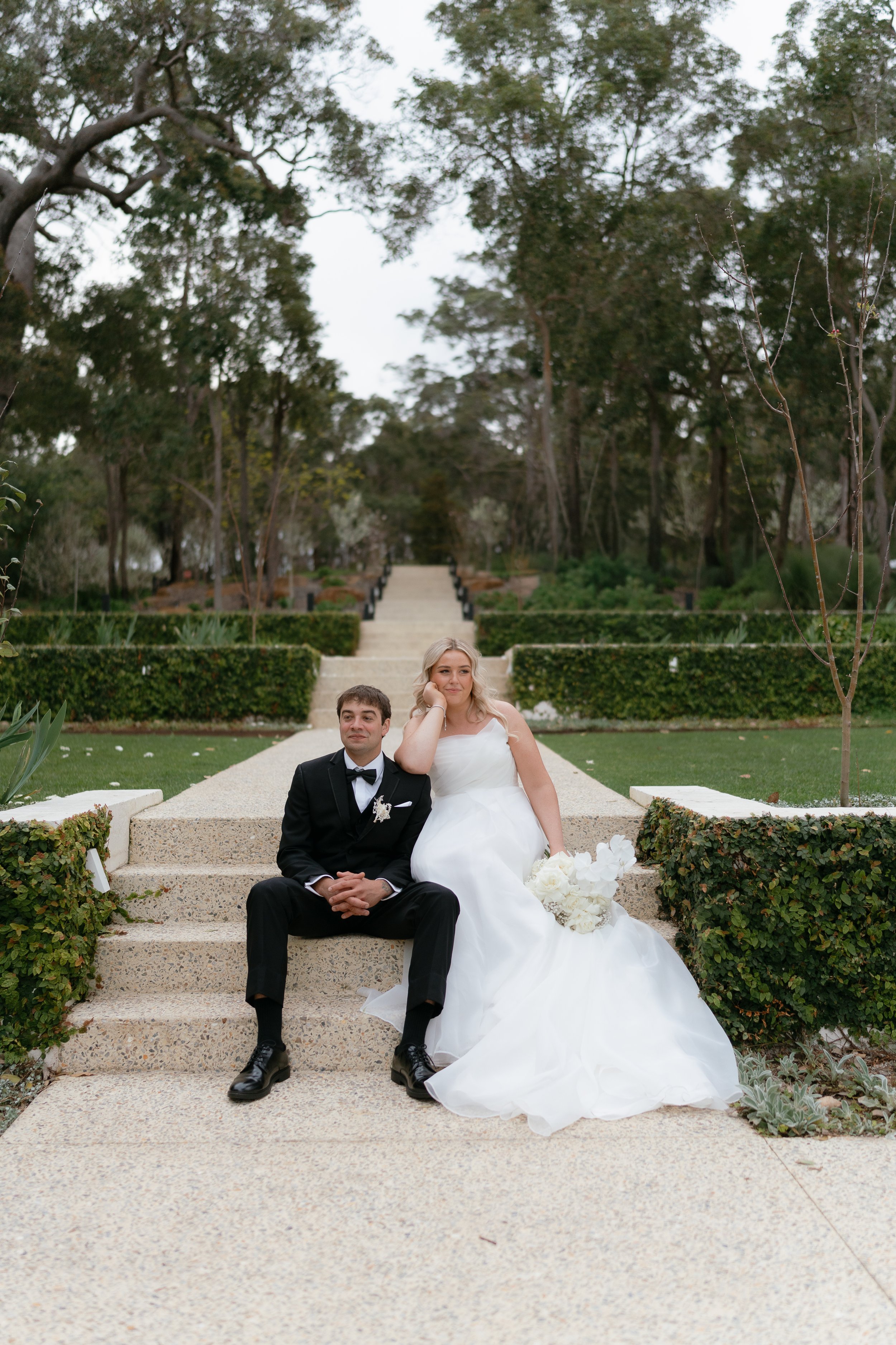A bride and groom sitting on outdoor steps surrounded by greenery.