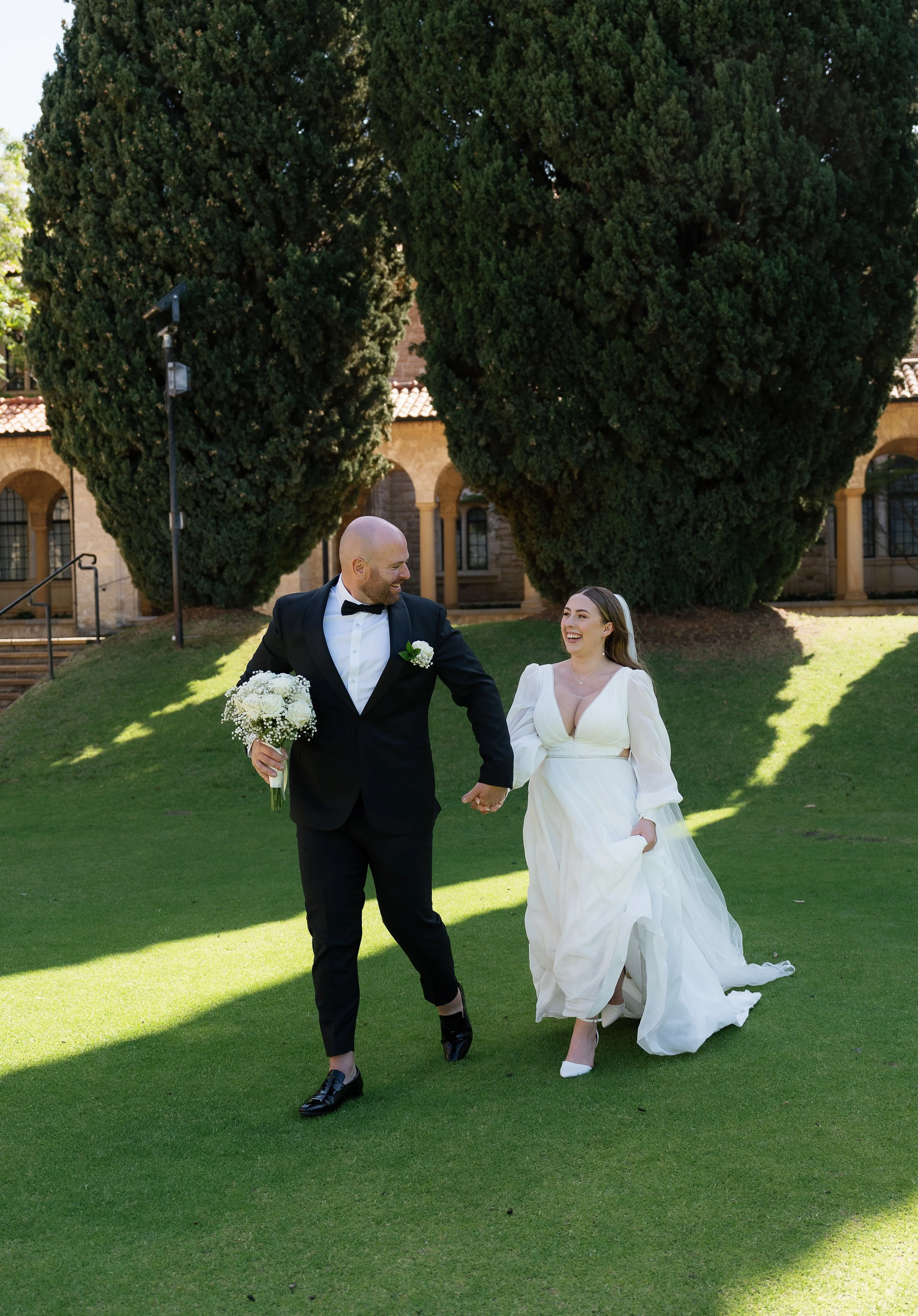 A bride and groom walking hand in hand outdoors on a sunny day, smiling at each other, with large trees and a historic building in the background.