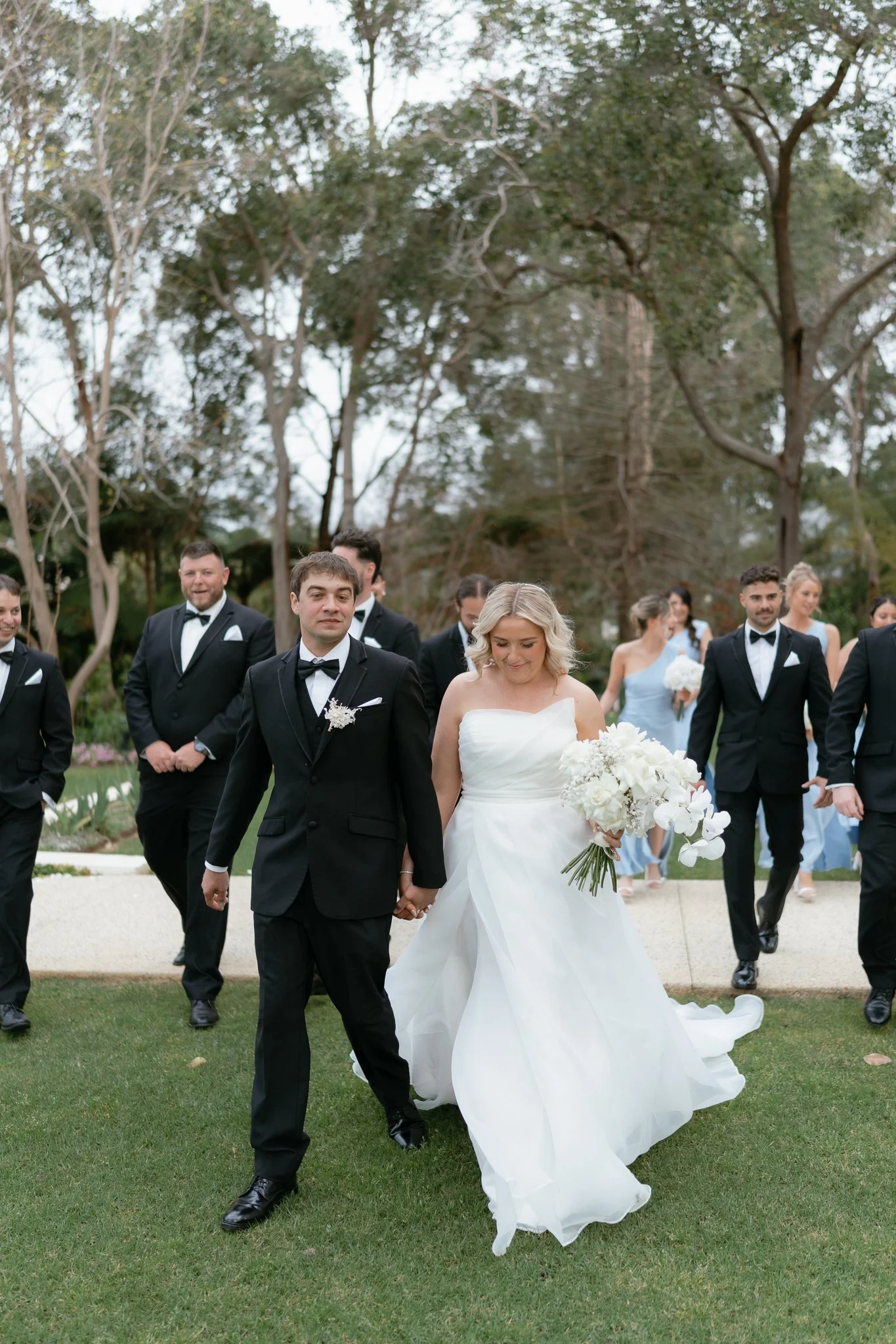 A bride in a white wedding dress holding a bouquet of white flowers walking outside, surrounded by groomsmen and bridesmaids in formal attire.