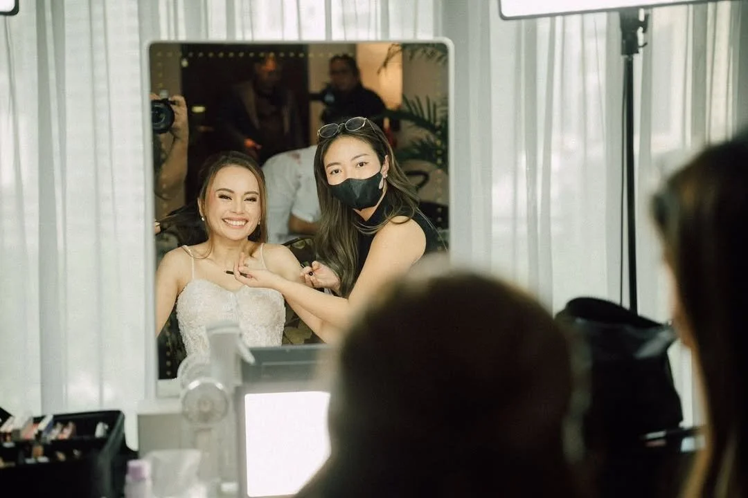 Bridal makeup artist applying makeup to a smiling bride in front of a mirror, with the artist wearing a face mask, in a well-lit room.