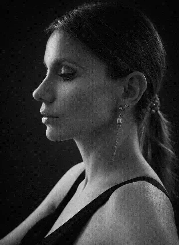Side profile of a woman with smooth hair tied back, wearing an earring and a dark sleeveless top against a dark background.