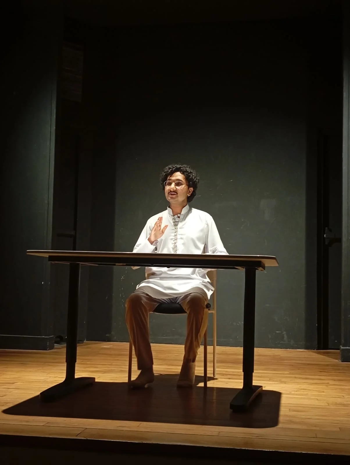 Person with dark curly hair, glasses, and mustache sitting at a desk on a stage, gesturing with one hand, in a dark room with black walls and a wooden floor.