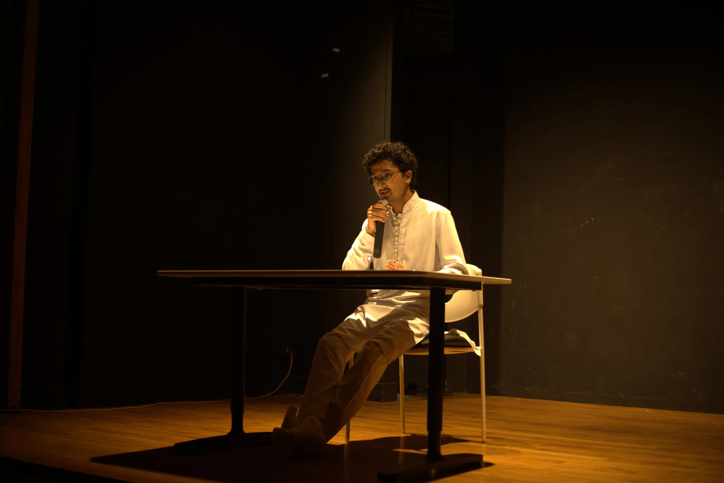 A man with glasses and curly hair, dressed in a white shirt and beige pants, sitting at a table on a stage, talking into a microphone with a spotlight on him.