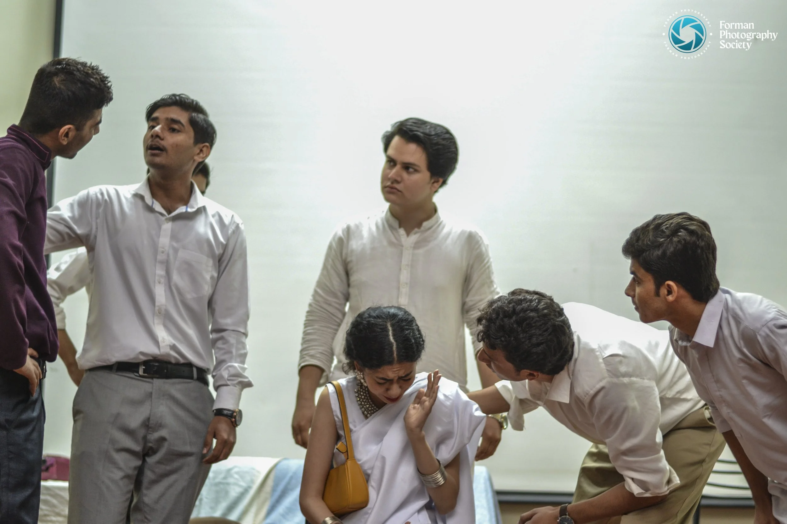 A group of six people in a room, one woman appears distressed and is being comforted by two men, while three other men are engaged in discussion around her.