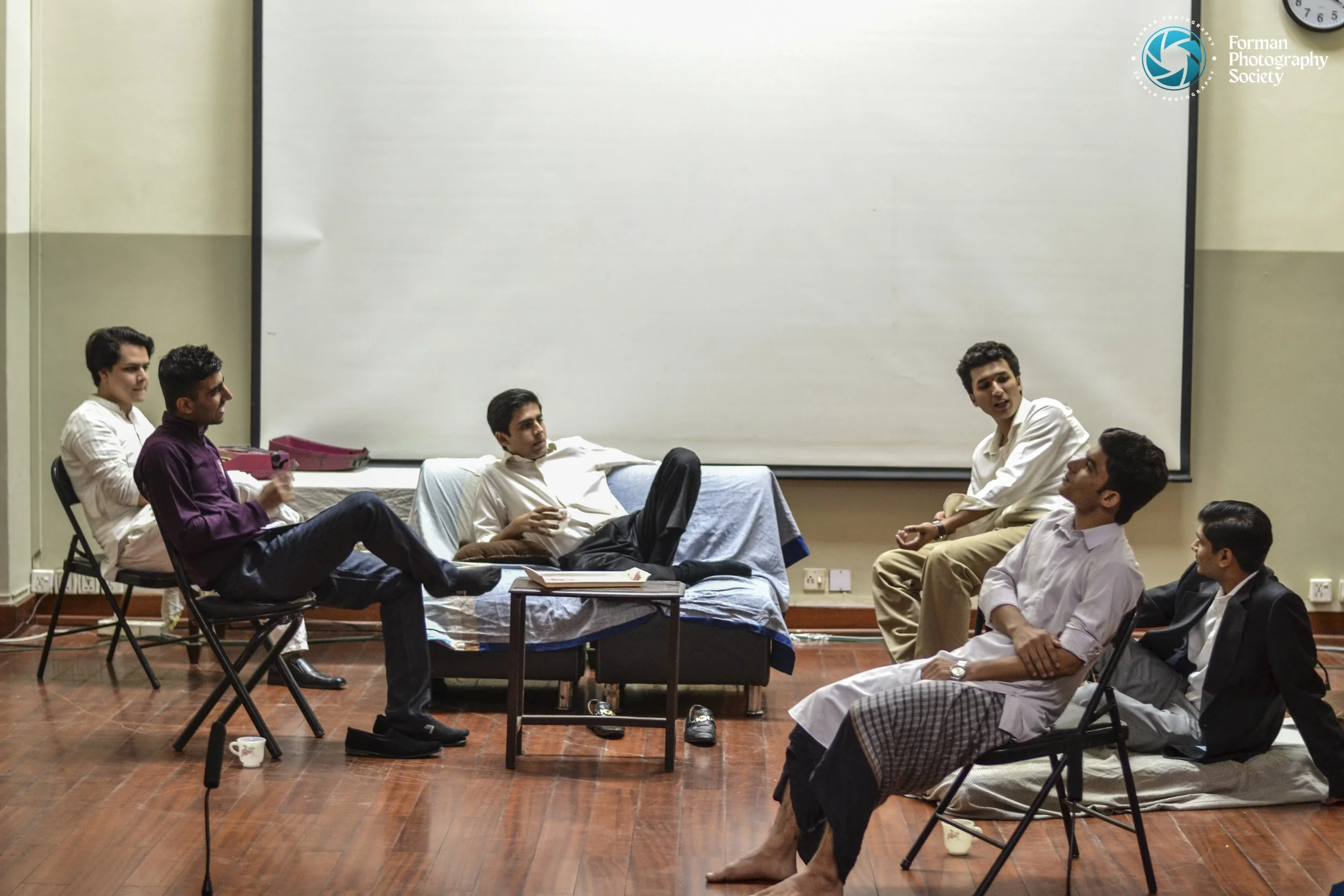 Group of seven young men in a classroom or meeting room, sitting on chairs and a sofa, engaged in a discussion or meeting, with a large whiteboard behind them.